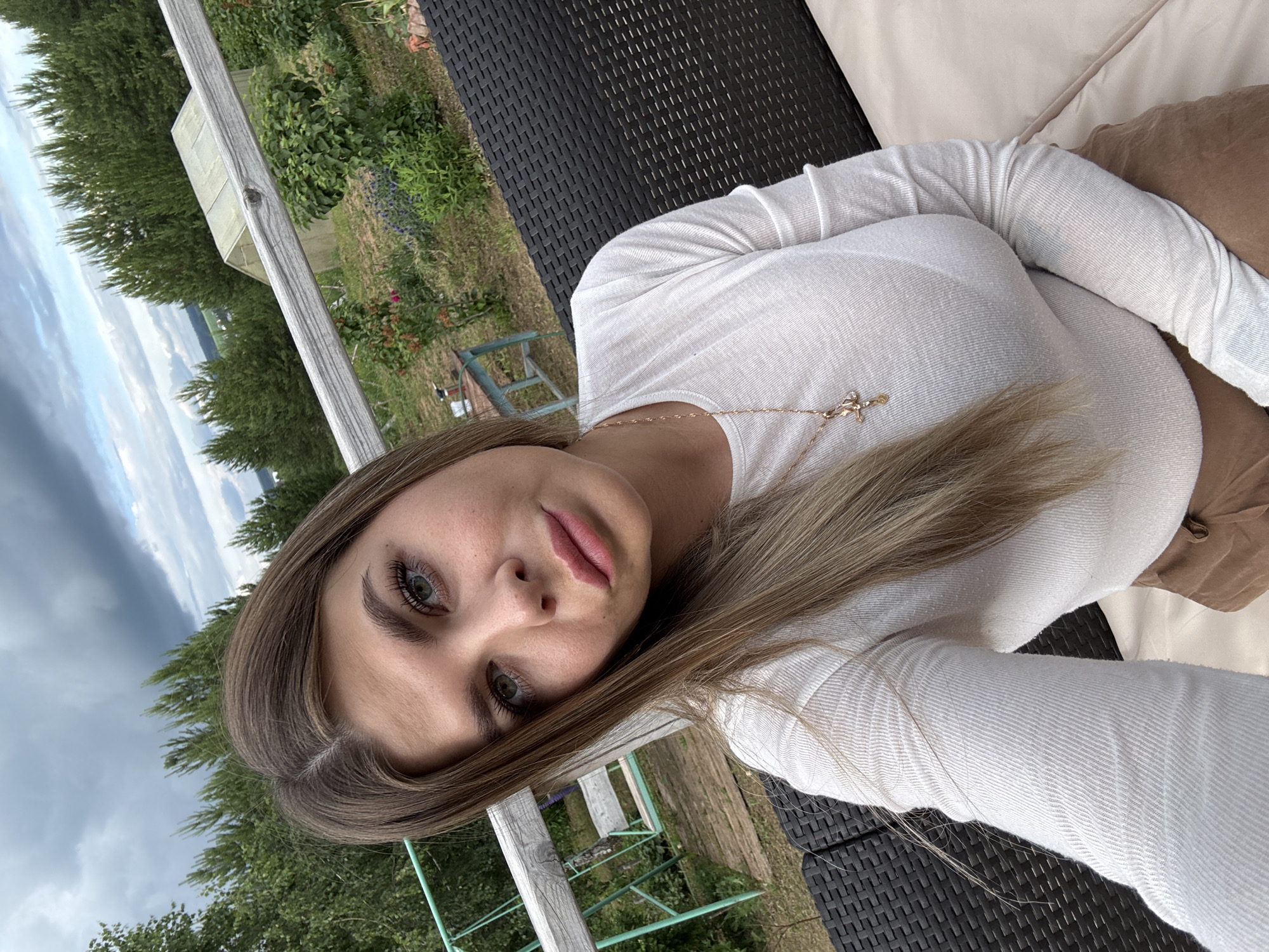 A young Caucasian woman with long, light brown hair is seated outdoors, likely in a rural or suburban setting. She is wearing a white, long-sleeved, fitted shirt and brown pants. A gold necklace with a cross pendant is visible. The background suggests a garden or yard with trees, a greenhouse, and a wooden fence. The sky is cloudy. The location is identified as Фоки, Russia.