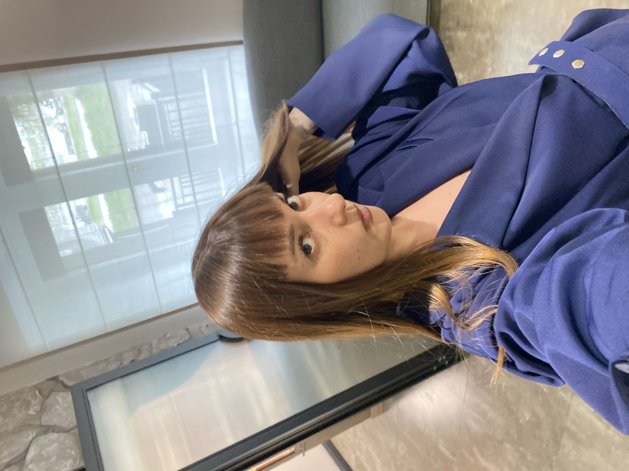 A woman with brown hair and bangs is lying on a gray couch, wearing a blue blazer. She is looking at the camera. Behind her, there is a window with white blinds. A black-framed glass door with a stone wall next to it is also visible. The location is possibly an interior space. The city of Votkinsk, Russia, cannot be determined from the image.