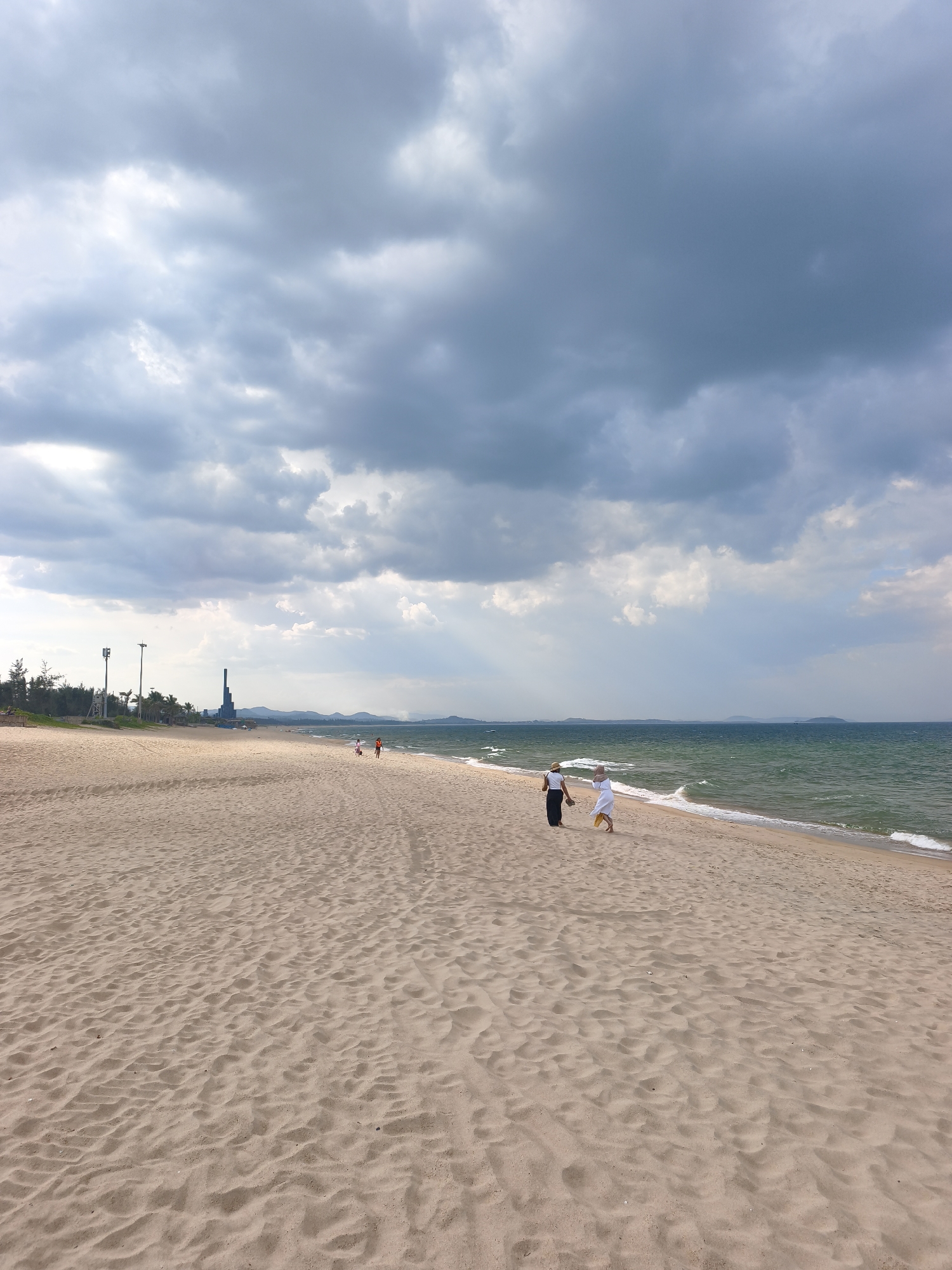 This is a photograph of Tuy Hoa beach in Vietnam. The image captures a wide expanse of sandy beach leading to the ocean. The sky is dramatic with dark, heavy clouds that contrast with patches of lighter sky. Rays of sunlight pierce through the clouds, illuminating parts of the scene.
In the foreground, the sand is textured with ripples, suggesting wind or water action. Further back on the beach, two individuals are walking towards the left, one in white and the other in dark clothing. Further in the distance, another person is walking. Along the coastline, palm trees and other vegetation are visible, bordering the sandy area. On the left side of the image, a tall, dark, industrial-looking structure stands against the horizon, possibly a power plant or refinery, along with some utility poles. The ocean water is a vibrant green, with moderate waves breaking on the shore. The horizon is visible, with distant landforms or islands appearing as faint shapes.