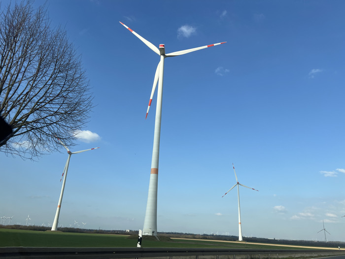 The image displays an expansive outdoor scene featuring a wind farm in Eschweiler, Germany, captured under clear daylight conditions. The sky is a deep blue with scattered white cumulus clouds. In the immediate foreground, a dark, indistinct object partially obscures the lower-left corner. A bare deciduous tree with fine, leafless branches occupies the left side of the frame. A dark guardrail extends horizontally across the lower third of the image, separating the foreground from a flat, green agricultural field. A white and black roadside marker is visible near the guardrail, positioned at the base of the central wind turbine.
Three prominent wind turbines dominate the midground. The largest turbine, centrally located, features a tall white cylindrical tower with an orange band encircling its lower section. From its nacelle, three white blades extend, each tipped with a red segment. The blades are static, with one pointing upward and two downward. To the left and slightly behind, a second, similar wind turbine is partially visible, also exhibiting a white tower, orange band, and red-tipped white blades. A third, smaller turbine is positioned to the right and further back, characterized by a white tower and red-tipped white blades. In the distant background, numerous additional wind turbines are scattered across the landscape, indicating an extensive wind farm. No individuals are visible, and no specific actions or interactions are discernible within the scene.