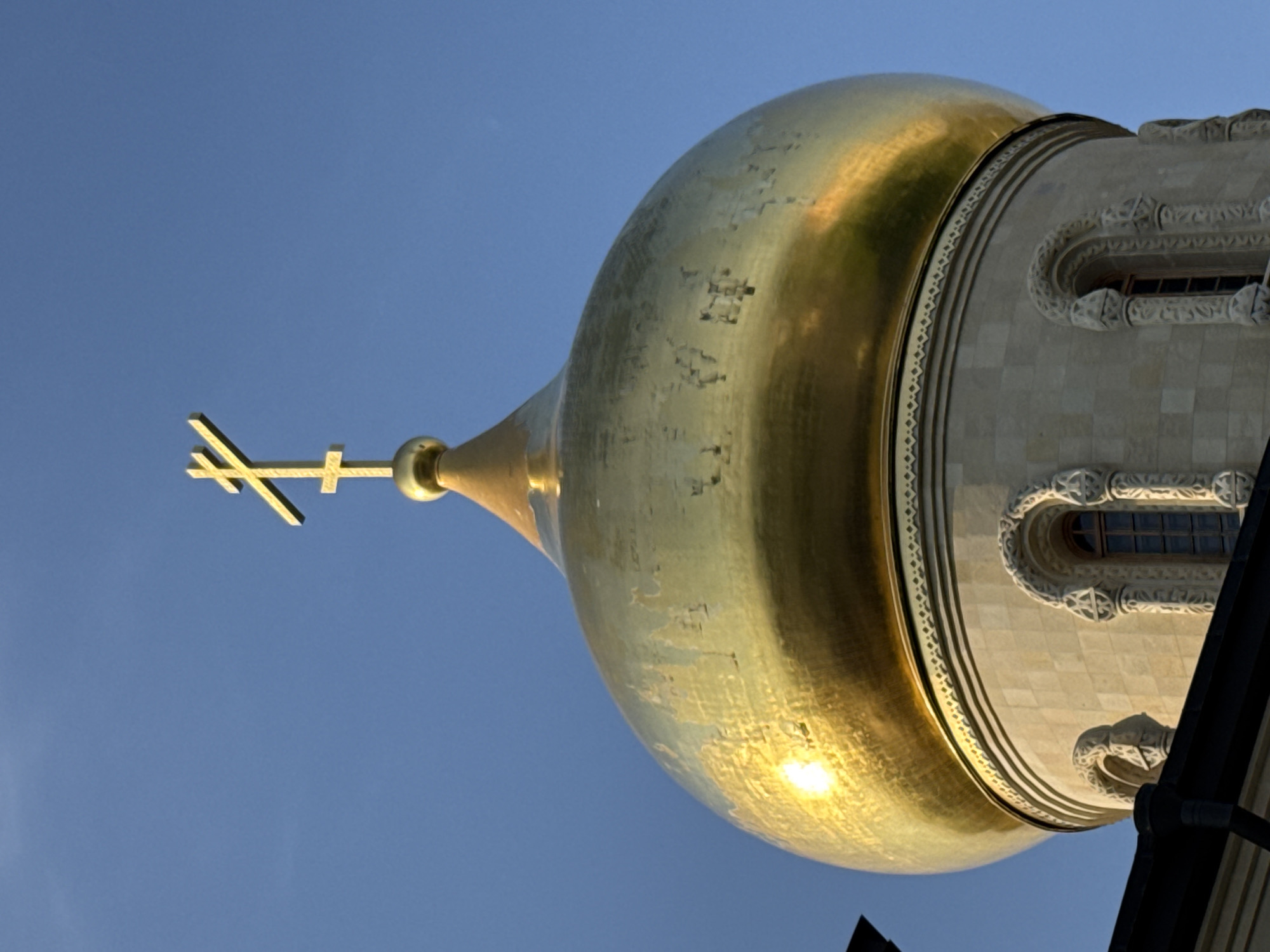 A golden Orthodox cross tops a golden dome of a building, likely a church, against a clear blue sky. The dome is bulbous and reflects sunlight intensely on its left side, showing some wear and tear. Below the dome, the building's stone facade features arched windows with ornate stonework. The image is a close-up, architectural photograph of religious iconography. The location is the city of Kirishi, Russia.