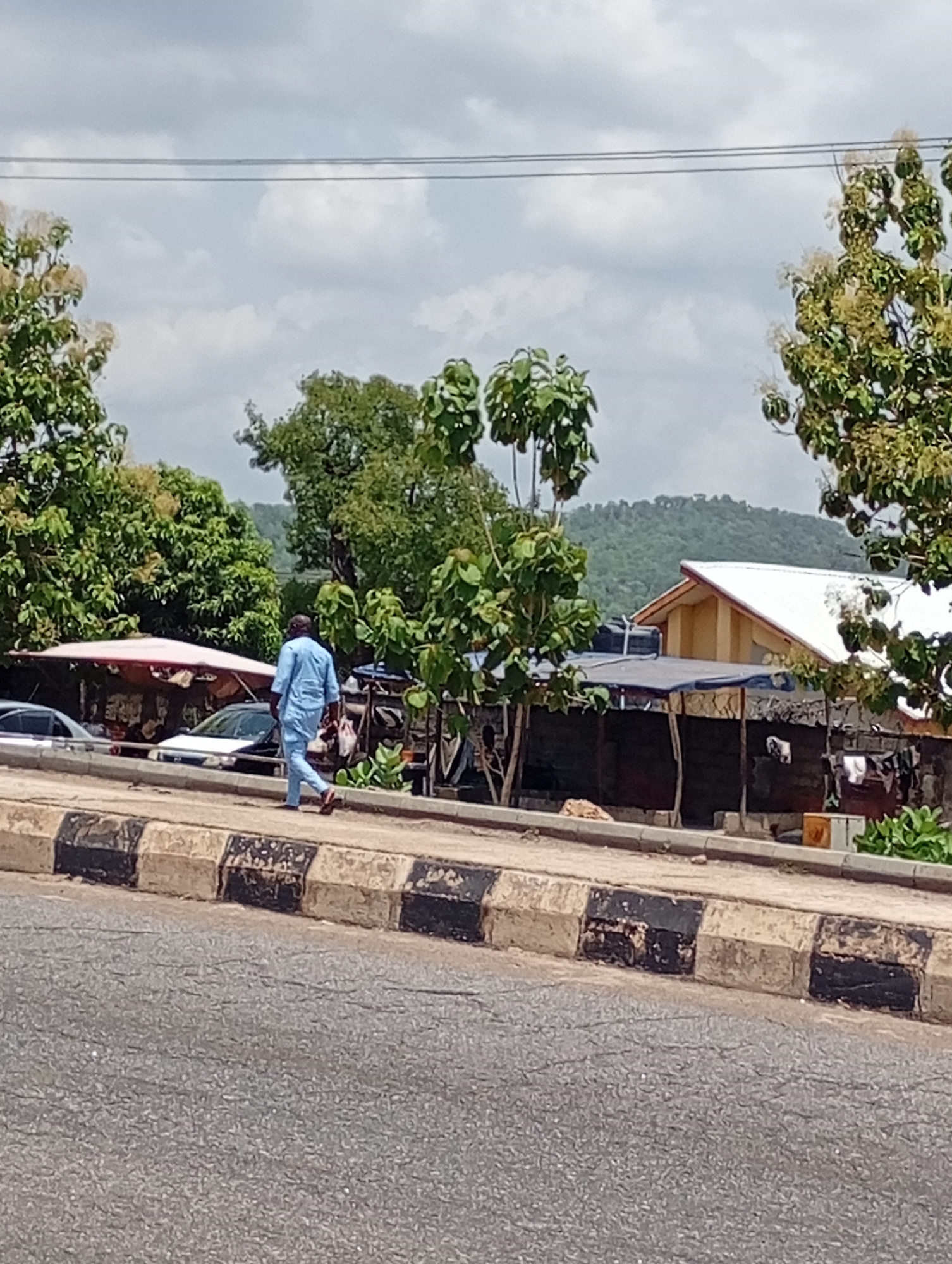 The image shows a man walking along a sidewalk in a city. The sidewalk is made of black and white blocks, and there is a road in the foreground. In the background, there are trees, buildings, and a hill. There are also parked cars, canopies, and clothes hanging out to dry. The sky is cloudy. The location is Unknown, Unknown.