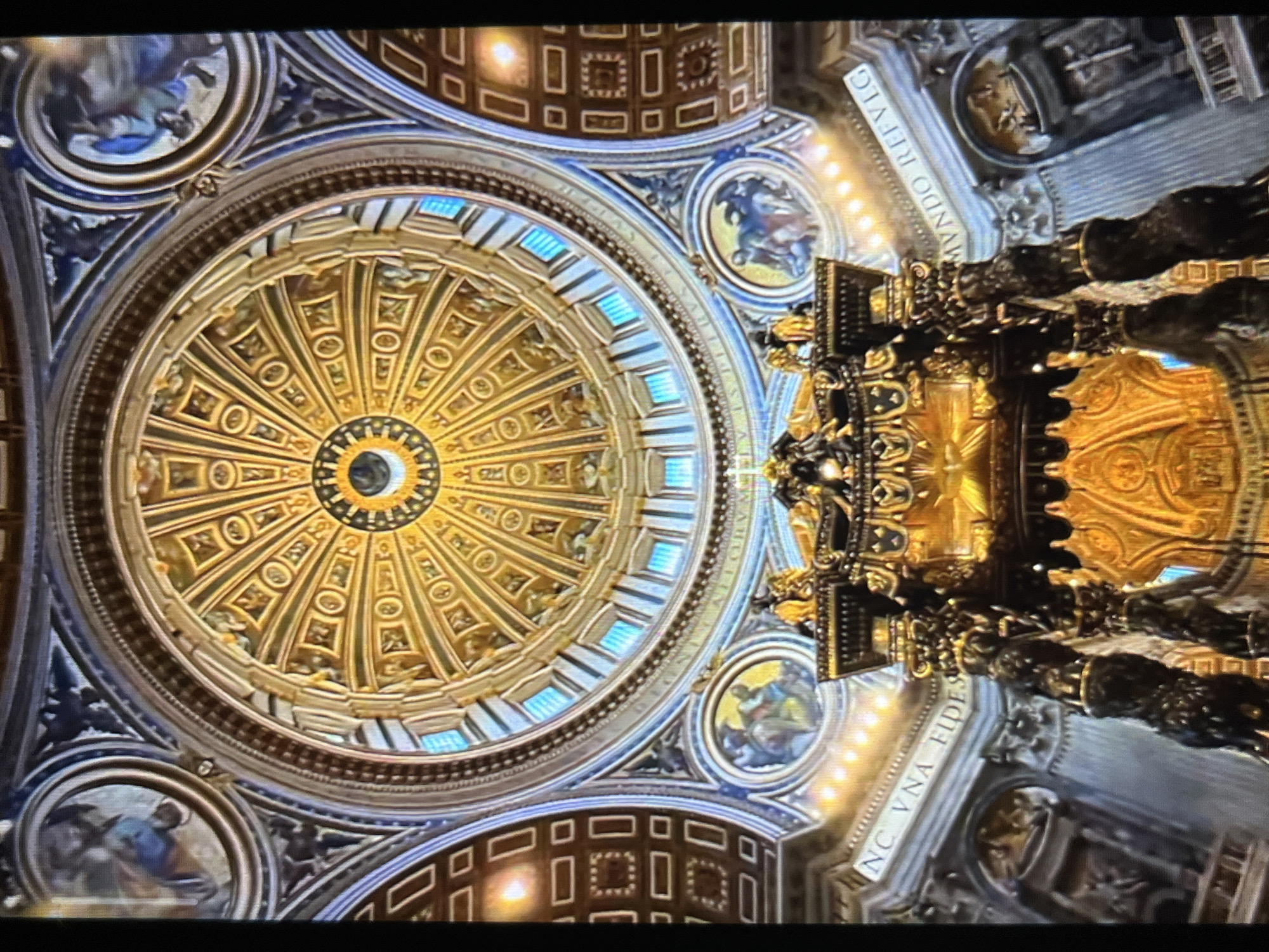 The image depicts the interior of a large, ornate dome, likely within a basilica or cathedral. The dome is decorated with intricate gold designs and radial patterns. There are circular depictions of people around the base of the dome, and a section featuring square patterns in a dark color. The view is angled upwards towards the center of the dome. Part of a complex, ornate structure, possibly a canopy or altar, appears in the lower right corner. The location is NOT Tinaquillo, Venezuela.