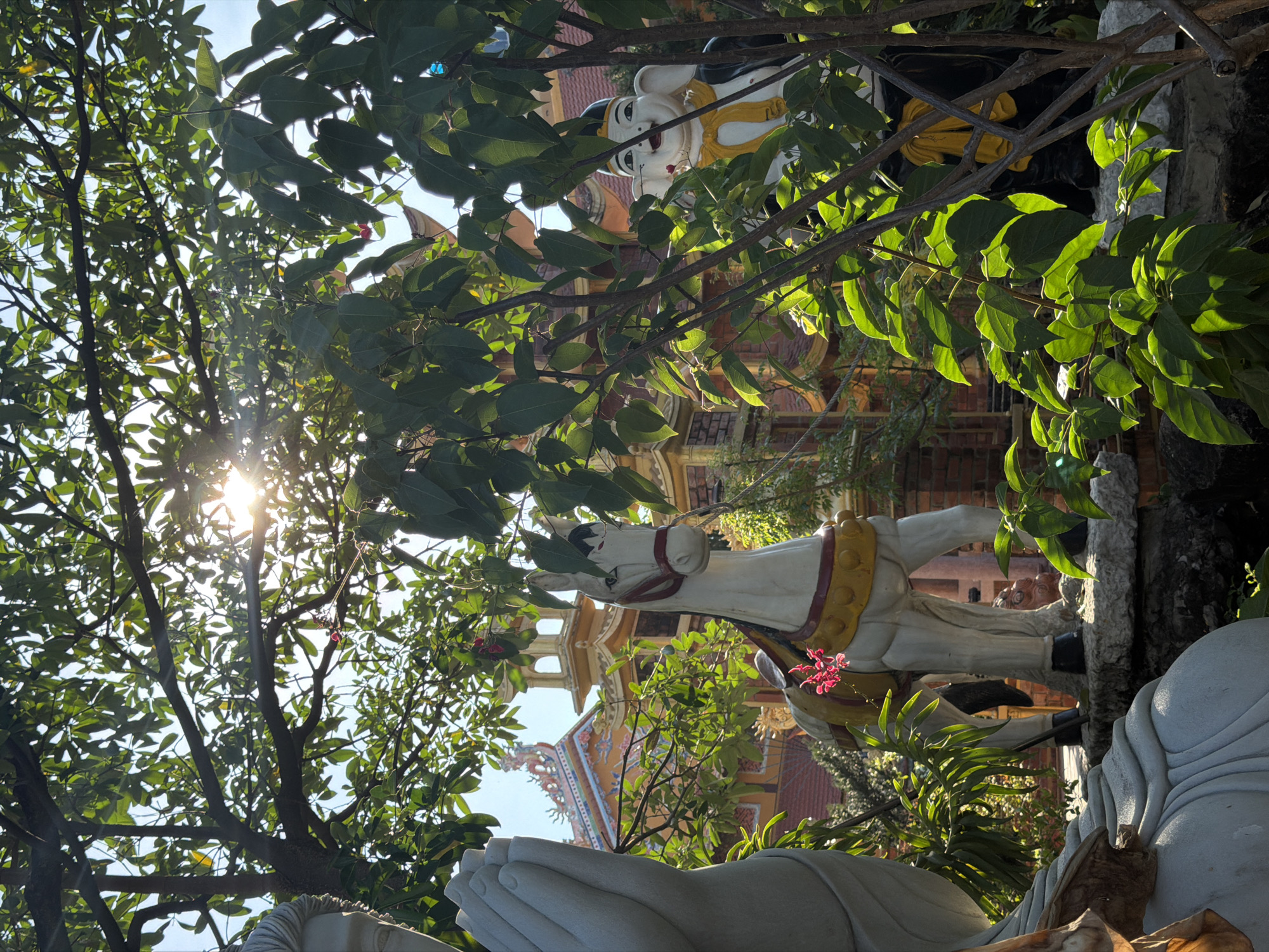 The image shows a scene at the Quan The Am Pagoda in Đà Nẵng, Vietnam. In the foreground, there is a portion of a large, white statue depicting two clasped hands. Behind this statue, a white horse statue with a yellow harness is visible amidst trees. Further back, a building with a reddish-brown facade and ornate roof decorations can be seen. Above, through the tree branches and leaves, a figure with a white face and yellow clothing is discernible, and the sun shines brightly. The photograph appears to be taken at an upward angle.