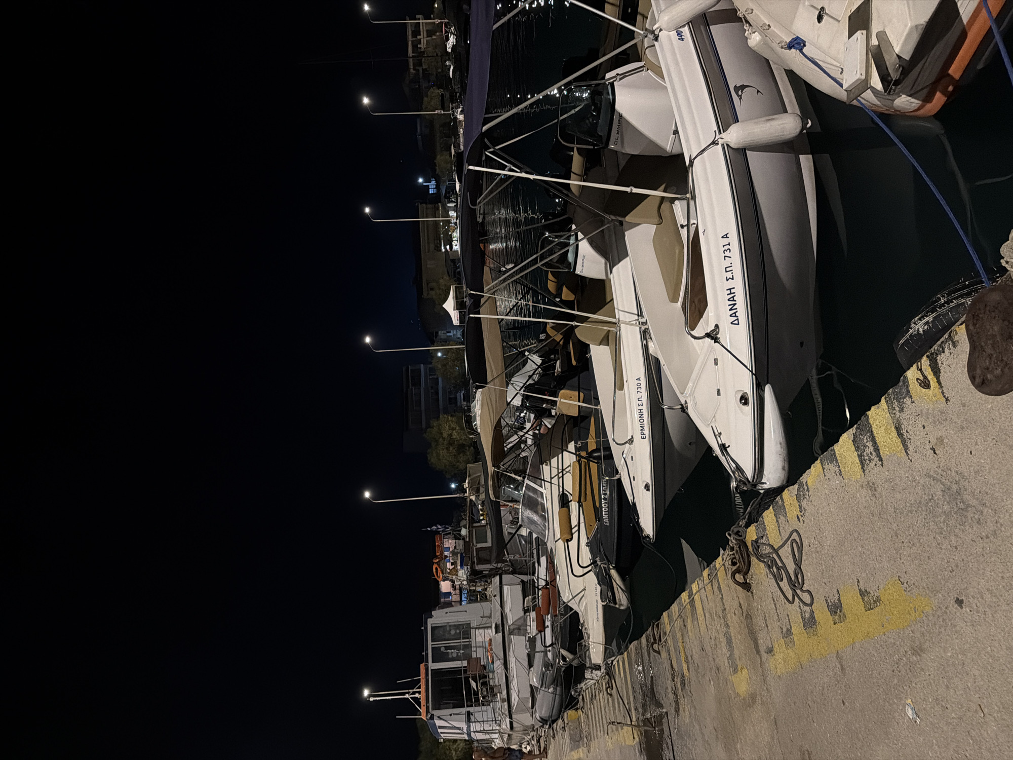At night, a row of boats is docked at a harbor. The boats are white with beige seating and dark blue canopies. Some of the boats have names written on their sides in Greek. There is a concrete dock with yellow markings, and chains are visible connecting the boats to the dock. In the background, there are buildings and streetlights. The water is dark and reflects the lights. This scene is located in the Municipal Unit of Alyzia, Greece.