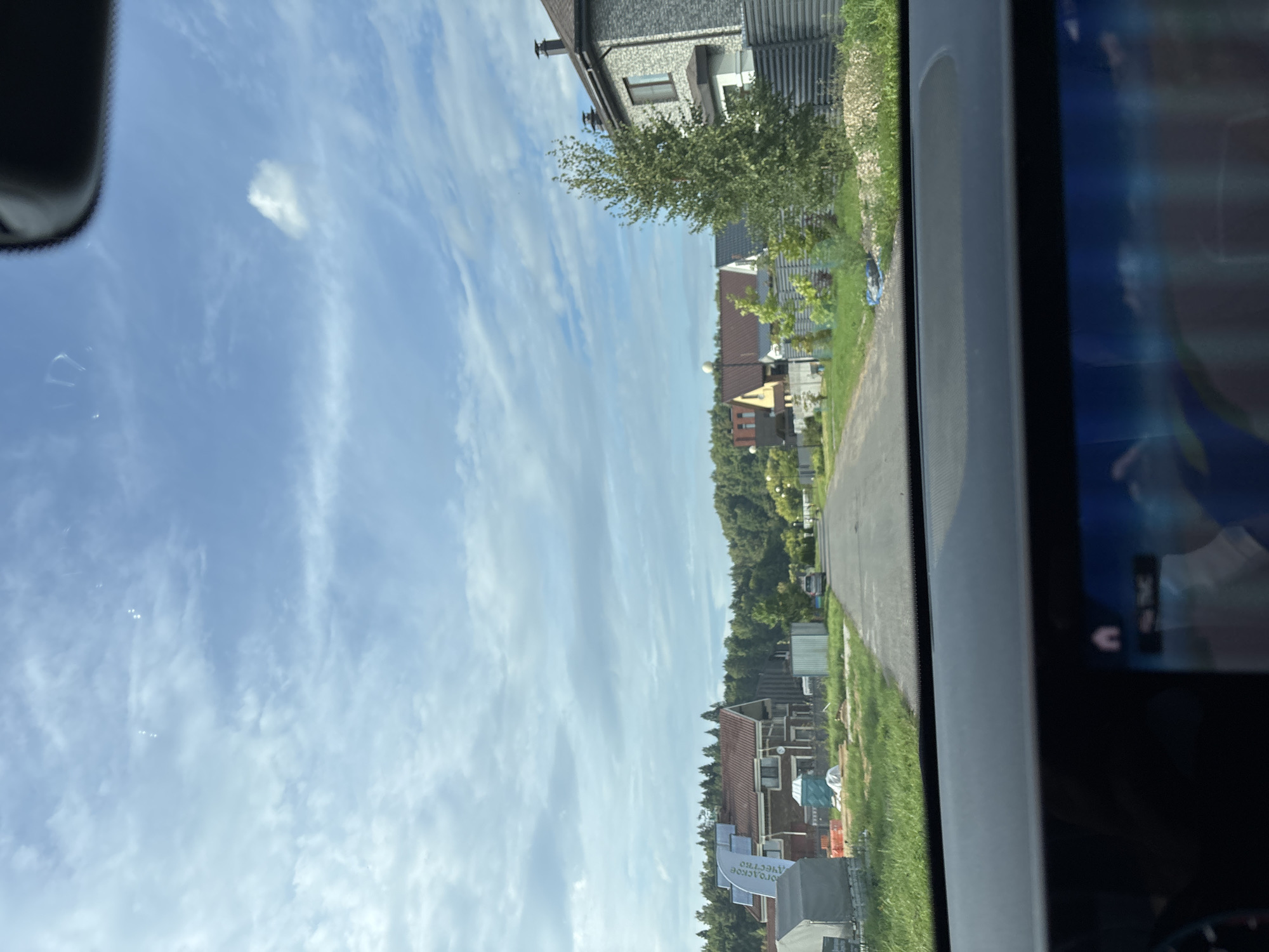 The image shows a view from inside a car, looking out onto a suburban street in Zvenigorod, Russia. The street is lined with modern houses, some of which appear to be under construction. Green trees and lawns surround the houses, and the sky is partly cloudy. A small, dark-colored car is visible driving away from the camera down the street. The dashboard of the car is visible in the foreground, with a navigation screen displaying a map.