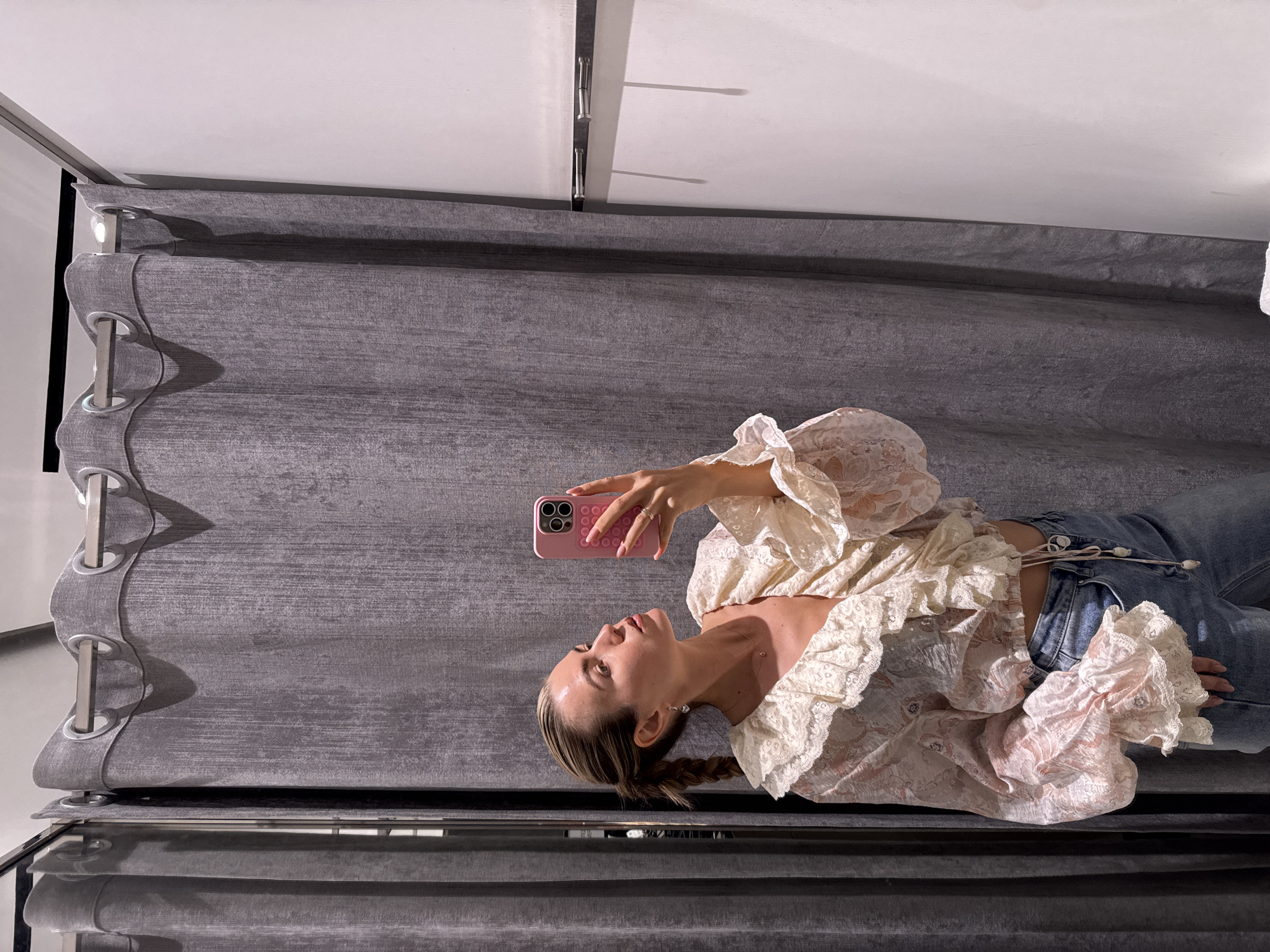 A woman with blonde hair styled in a braid is lying on a gray, upholstered surface, possibly a couch or bench, indoors. She is wearing a white, lacey top and blue jeans. She holds a pink smartphone in her hand, seemingly taking a selfie. Overhead, gray curtains hang from a track system on a white ceiling. The exact location in an unknown city is not discernable from the image.