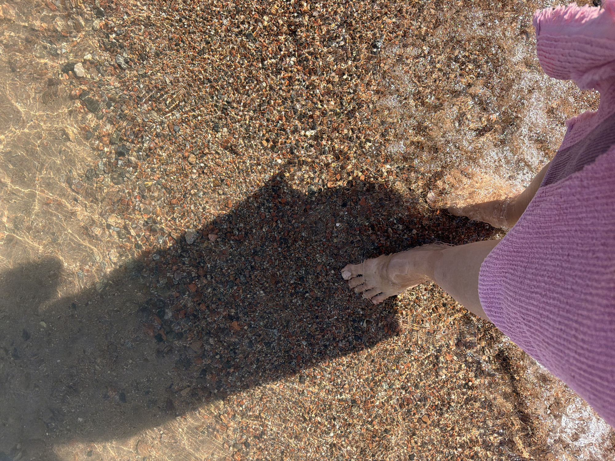 The image shows a person's feet walking in shallow, clear water over a pebble beach. The water is transparent, revealing the textured seabed below. Light refracts through the water, creating shimmering patterns on the pebbles. A large shadow, presumably from the person taking the photo, stretches across the left side of the frame. The person is wearing a pink garment, partially visible in the foreground. The scene suggests a recreational activity at a beach, likely during daylight hours. The location is specified as Choktal, Kyrgyzstan.