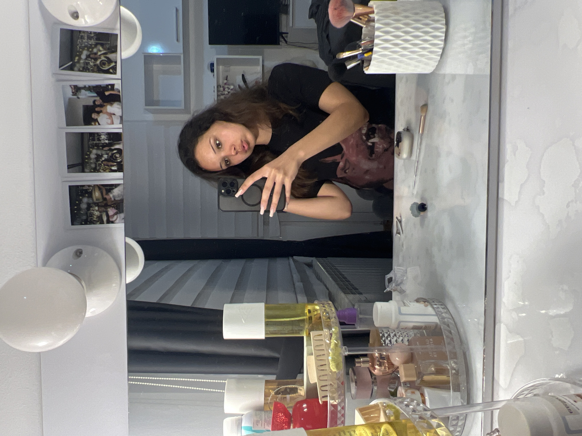 The image, taken in Zlatograd, Bulgaria, shows a young woman in her bedroom, reflected in a vanity mirror. She is holding her phone and taking a selfie, her facial expression is a pout. The room is decorated with several framed photos of people, and the vanity itself is cluttered with various cosmetic products, including perfumes, makeup, and brushes. The lighting appears to be artificial, coming from overhead fixtures.