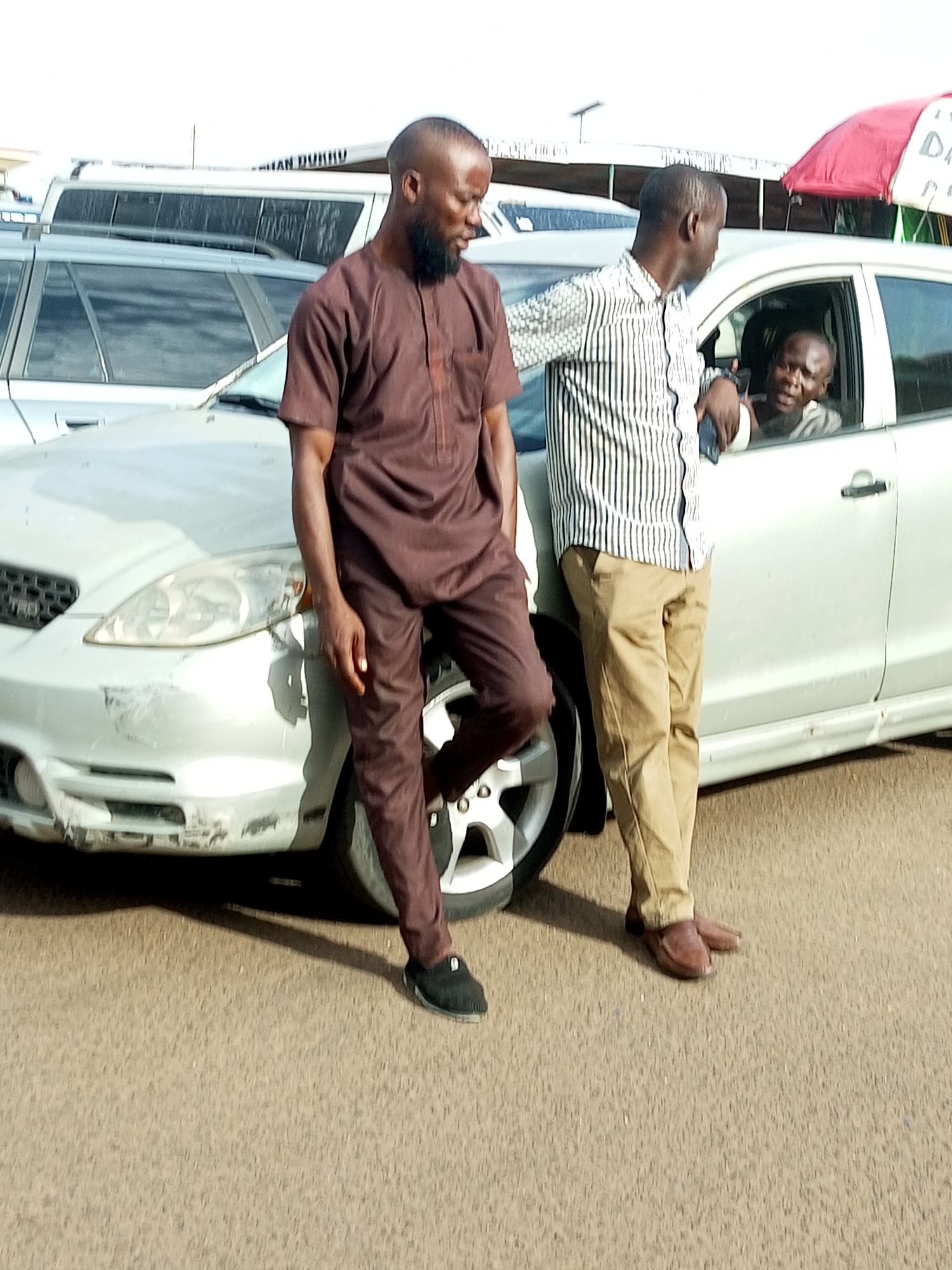 In Gombe, Nigeria, two men stand beside a silver sedan, one in a brown traditional outfit and the other in a striped shirt and tan pants. A third man is visible inside the car, looking out the driver's side window. The car appears to be parked on a paved surface, possibly a street or parking lot, with other vehicles and structures visible in the background. The scene suggests a casual encounter or a moment of pause.