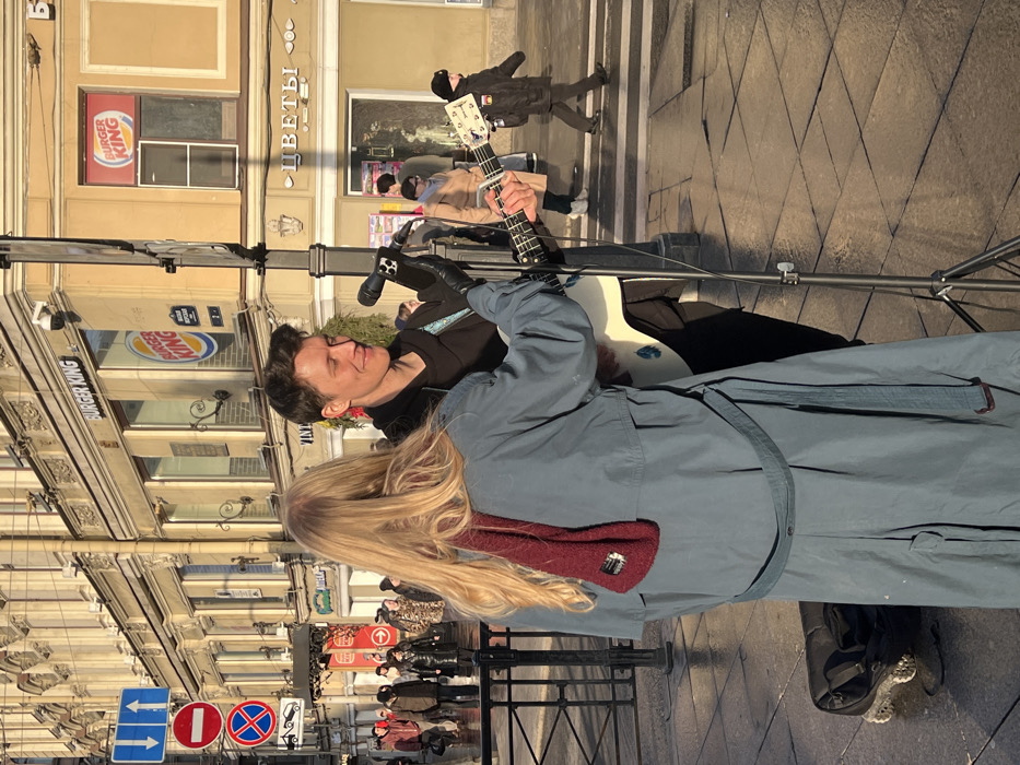 The image captures an outdoor street scene in Saint Petersburg, Russia, under bright, low-angle sunlight. In the foreground, two individuals are present. On the left, a person with long, wavy blonde hair, wearing a light blue or grey trench coat with a dark red knitted scarf, stands with their back mostly to the camera. Their right arm extends forward towards a male musician on the right. This male, with short dark hair, wears a dark hooded jacket and black gloves, and is seen smiling while playing a white acoustic-electric guitar. A microphone on a black stand is positioned in front of him, with an iPhone clipped to the stand. A black guitar case or bag is visible on the patterned tiled pavement to the left of the blonde-haired individual.
The background features a multi-story building with ornate architectural details, housing commercial establishments. Prominently visible are multiple "Burger King" logos and a shop sign in Cyrillic reading "ЦВЕТЫ" (Flowers). To the left, a pole displays several traffic signs, including a red circular "No Entry" sign, a blue circular "No Stopping" sign, and a blue square "Two Way Traffic" sign. Numerous pedestrians are walking in the background on what appears to be a wet street or sidewalk, indicating an urban environment.