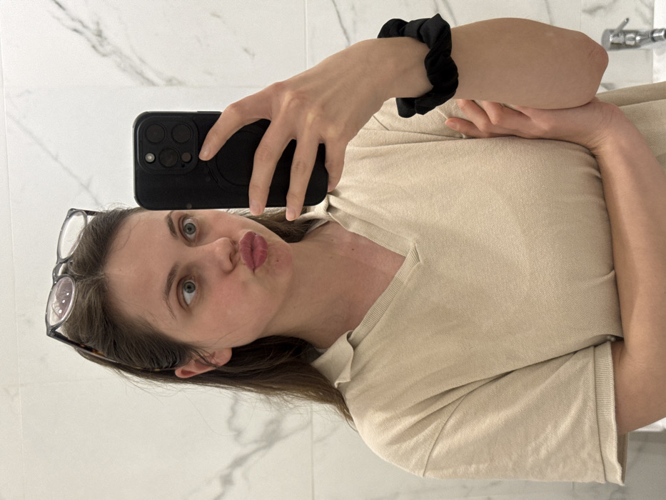 A young adult female is depicted in a mirror selfie, located in an interior space within Scafati, Italy, likely a bathroom or vanity area. She is shown from the upper chest upward, possessing medium-brown hair with dark-rimmed eyeglasses resting on her head. Her facial expression includes wide-open eyes, directed to her left, and lips pursed in a "kiss" formation. She wears a light beige or off-white short-sleeved V-neck top. Her left hand holds a dark-colored smartphone, visible from the back with a multi-lens camera array, positioned to capture her reflection. A black scrunchie is present on her right wrist. The background features white and grey marble-patterned wall tiles, and a metallic faucet fixture is partially visible in the lower right.