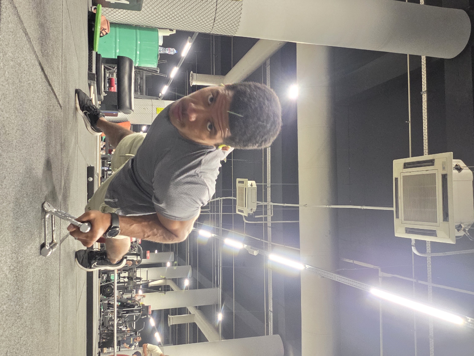 The image depicts a man performing a push-up exercise in a gym. He is positioned low to the ground, with his hands gripping metal handles that are resting on the floor. The man is wearing a gray t-shirt, light-colored shorts, and black athletic shoes. His facial expression is focused, and he appears to be mid-rep.
The environment suggests an indoor fitness facility. The floor is covered with dark, textured gym flooring. In the background, there are various pieces of gym equipment, including weight machines and what appears to be a vaulting horse. Lighting fixtures and an air conditioning unit are visible on the ceiling. The overall ambiance is that of a functional, well-lit gym. The image does not provide any specific visual cues that would allow for its location to be narrowed down to a particular city or country, including Varna, Bulgaria.