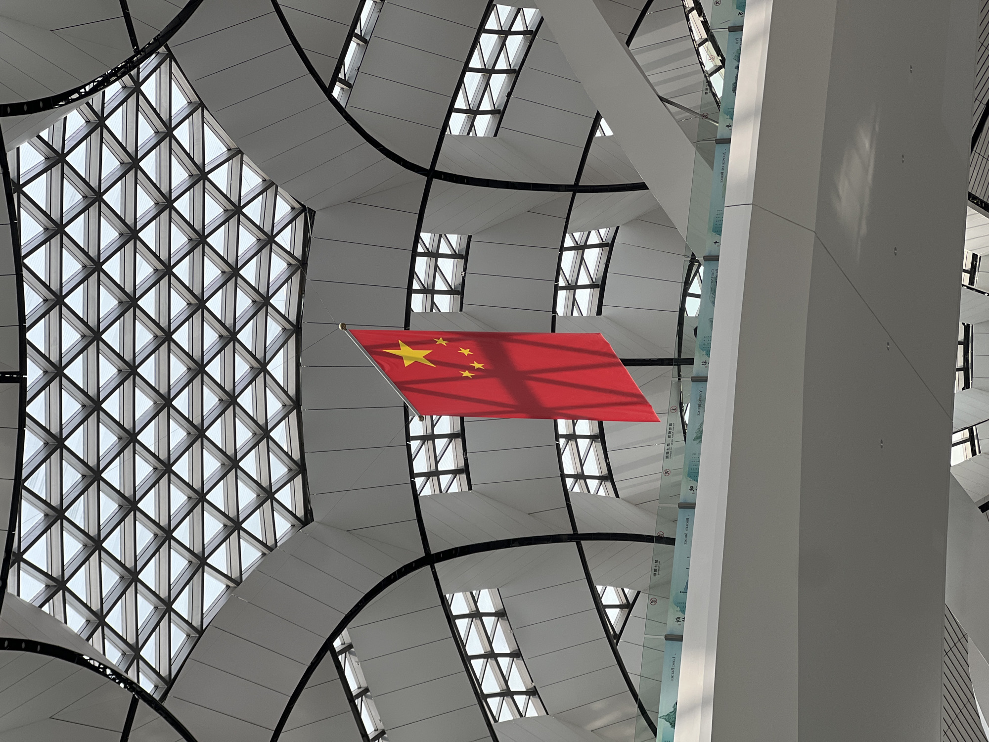 A low-angle, eye-level, color photograph shows the interior of the Beijing Daxing International Airport in Langfang, China, with a red Chinese flag prominently displayed.
The flag is in the center of the frame, with five yellow stars on the upper left corner. The flag is slightly tilted and appears to be suspended in mid-air.
The background is the interior architecture of the airport, featuring a complex geometric ceiling with diamond-shaped windows and white panels with black structural elements. A large grid of triangle glass panes is on the left.
A thick, white, angular column is on the right side of the frame. Glazed panels with Chinese text are attached to the column.