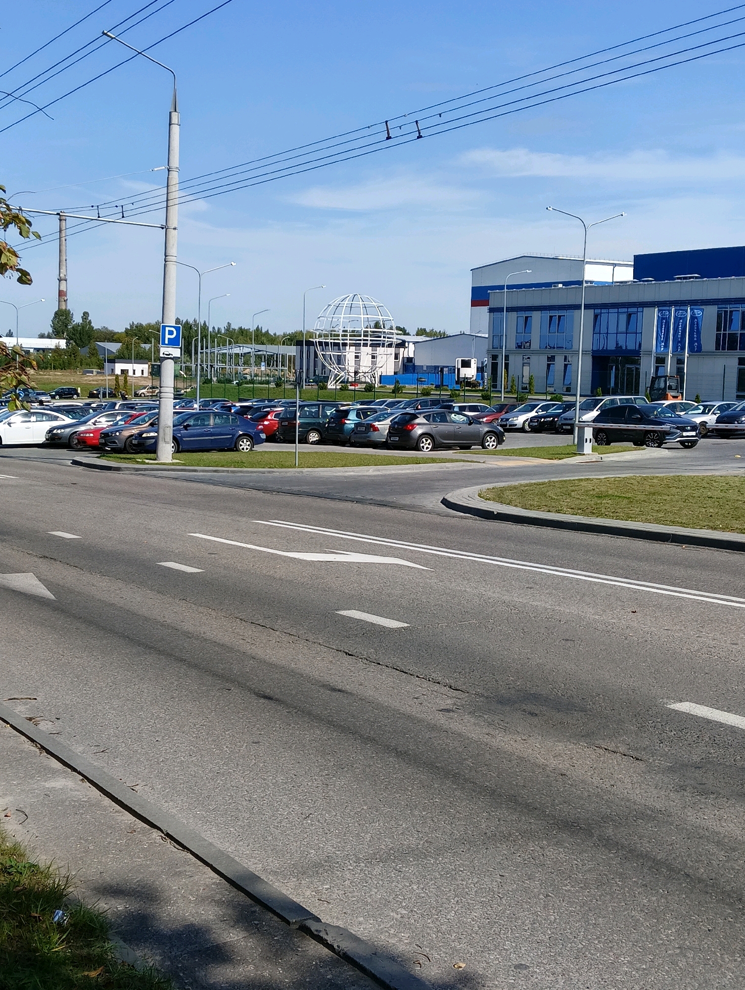 The image shows a parking lot filled with cars, a road in the foreground, and a modern industrial-style building in the background. The parking lot is situated in front of a long, blue and white building. Several cars are parked in rows, with a white SUV visible in the foreground of the parking lot. A large, white globe-shaped structure is also present in the parking lot, behind the parked cars and to the left of the building. The road in the foreground has white lane markings. A tall lamppost with a "P" parking sign is visible next to the road. Power lines crisscross the sky. In the distance, there are trees and a smokestack. The overall environment appears to be an industrial or commercial area.