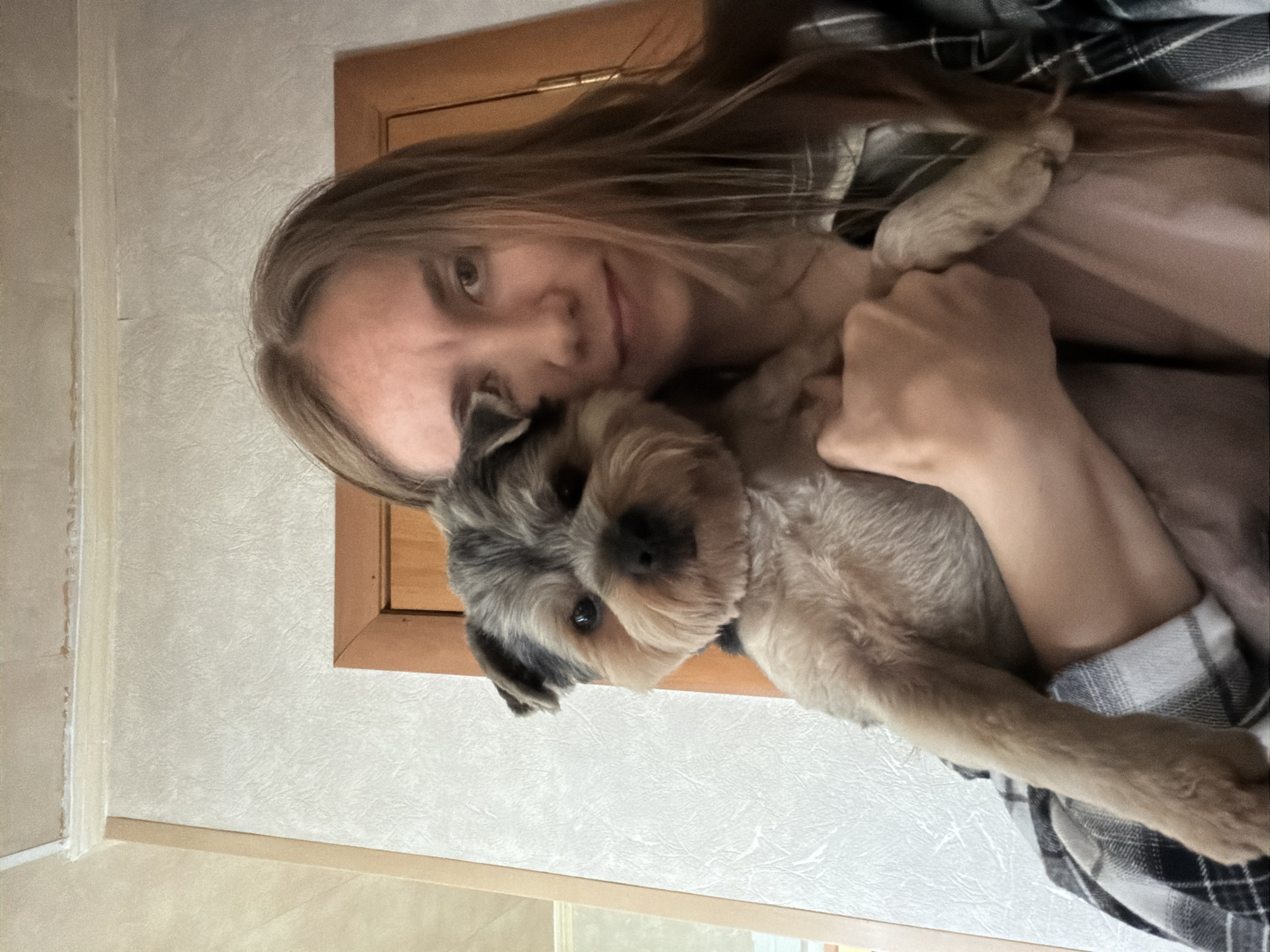 A young woman with long brown hair holds a small Yorkshire Terrier in her arms, looking directly at the camera. She is wearing a plaid shirt over a pink t-shirt. The background consists of textured wallpaper and a wooden doorframe. The scene appears to be indoors, possibly in a residential setting in Tambov, Russia.