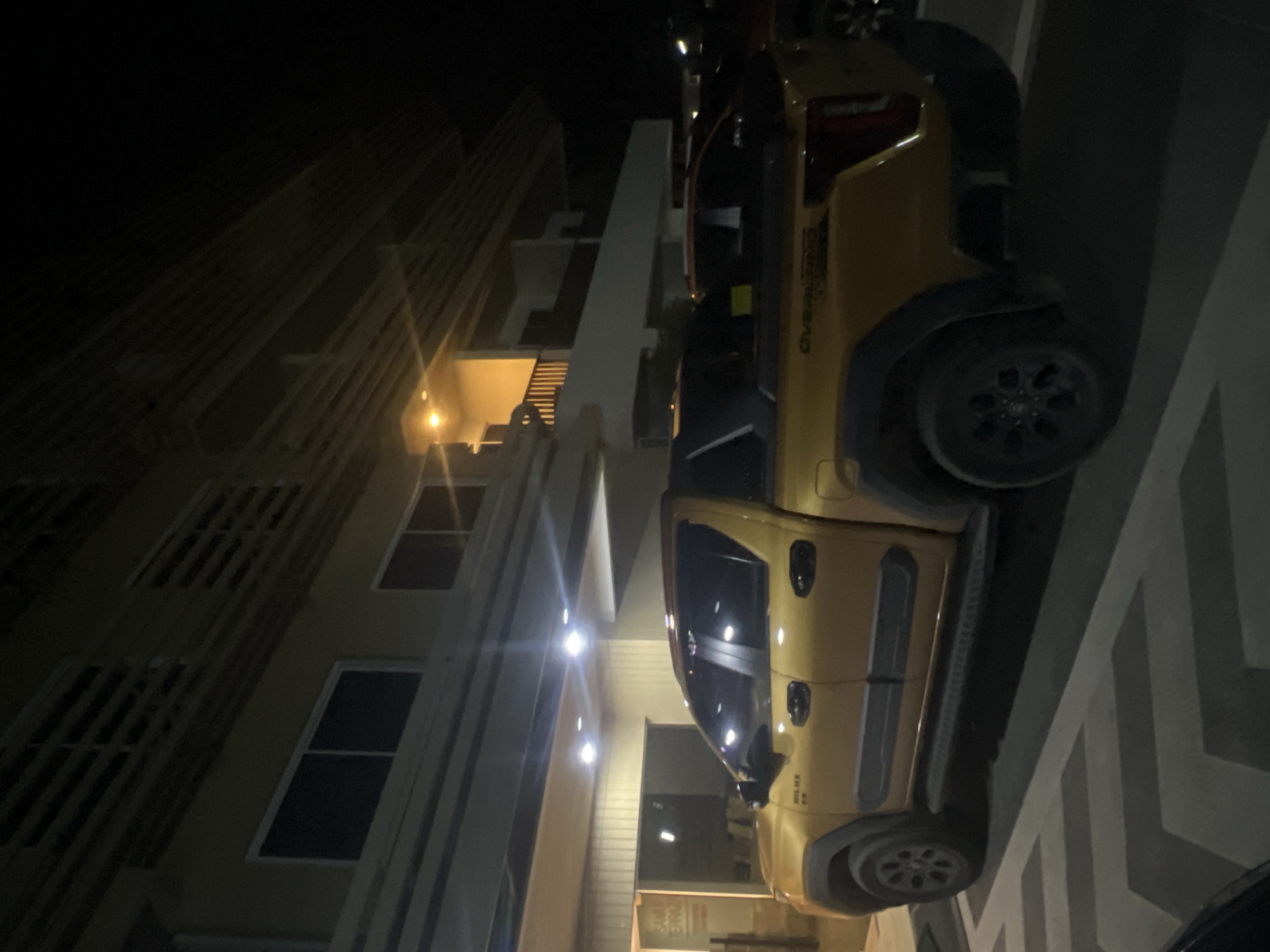 A yellow pickup truck is parked in front of a building at night. The truck is a double cab model with a silver stripe on the side. The building appears to be a hotel or apartment complex, with balconies and windows visible. There is a yellow sign on the side of the building that reads "SAMU TERRACES." The parking lot is paved with gray and white stripes. The scene is illuminated by artificial lights.