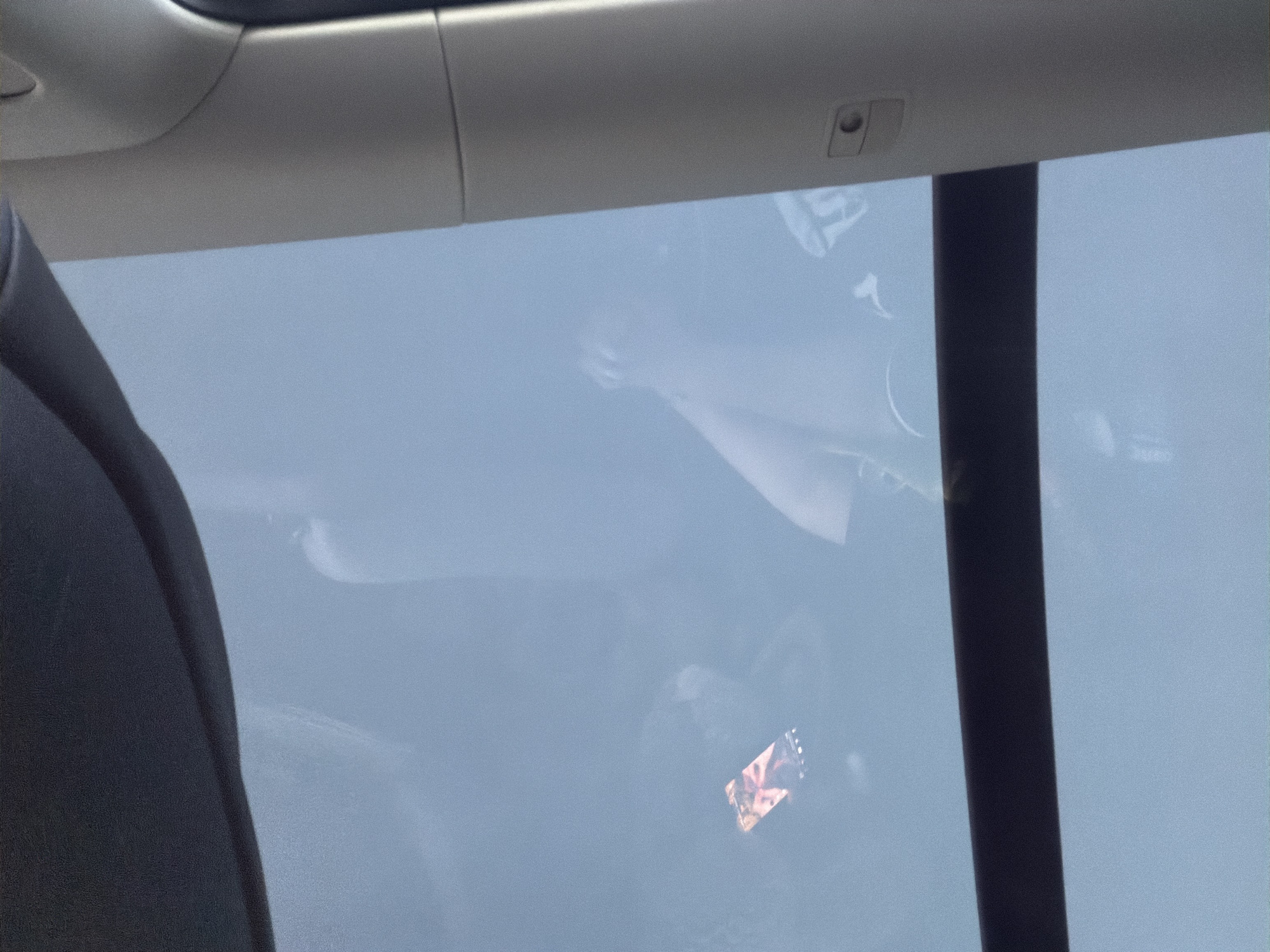 The image, taken in Dingle, Sweden, offers a view through a sunroof or panoramic roof of a vehicle. The sky appears overcast, casting a diffused light. A person is visible, their reflection or image subtly present on the glass. One hand is raised, and appears to be holding a smartphone. The phone screen displays a colorful image, possibly a photo or a graphic. The interior of the vehicle is also partially visible, including a dark trim along the top edge and a light-colored pillar on the right side, which has a small button or control on it. The overall impression is of someone using their phone while inside a vehicle, possibly on a journey.
