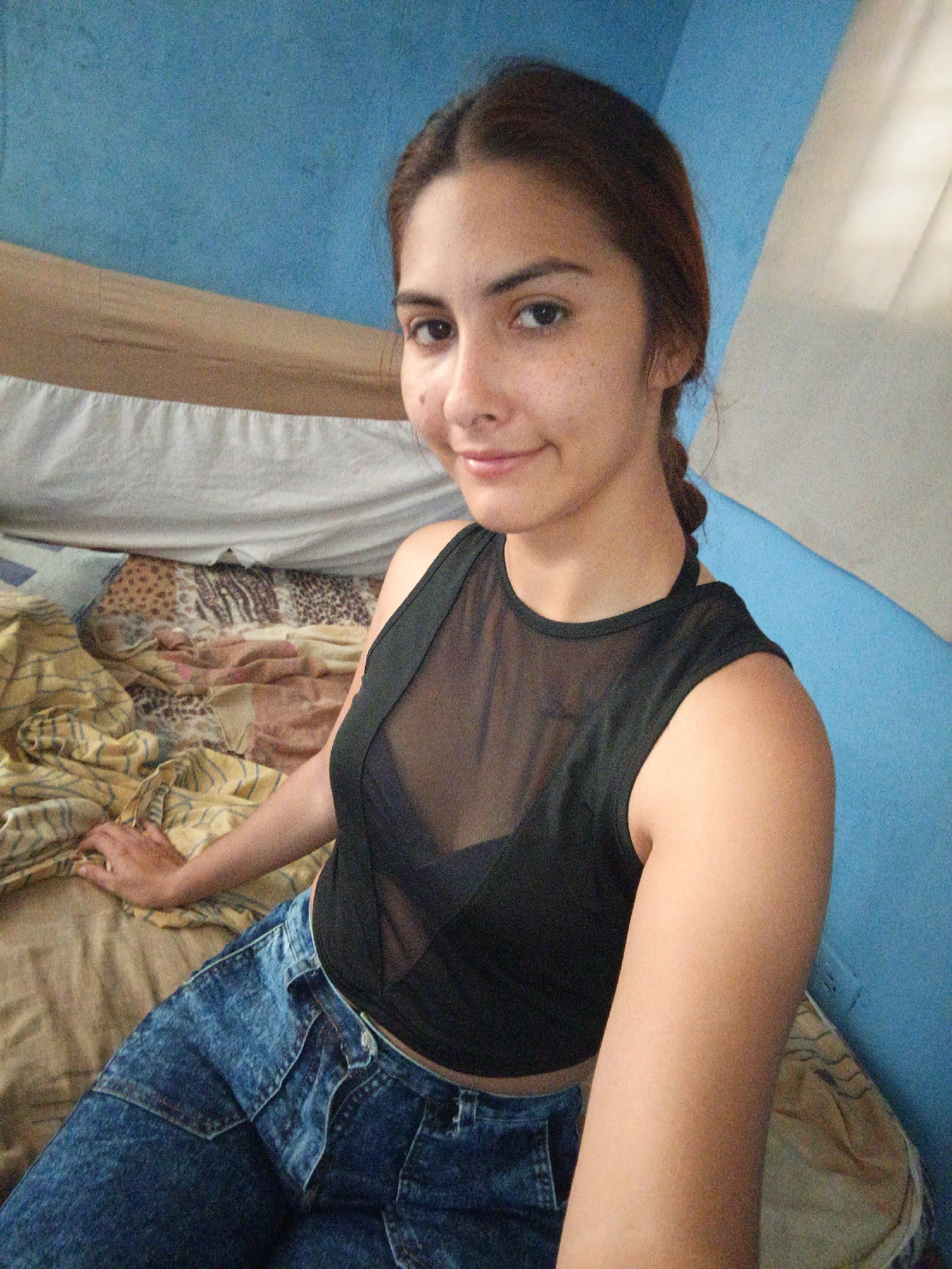 A young woman with dark hair in a braid is taking a selfie while sitting on a bed. She is wearing a black sheer top and blue denim jeans. The room has blue walls and a white curtain. The bed is covered with a patterned blanket and yellow sheets. The woman is smiling and looking at the camera. The image appears to have been taken during the daytime, as indicated by the natural light coming from the window. The setting suggests an indoor environment, possibly a bedroom in a home. The woman's expression and pose convey a casual and relaxed mood. There is no visible text in the image.