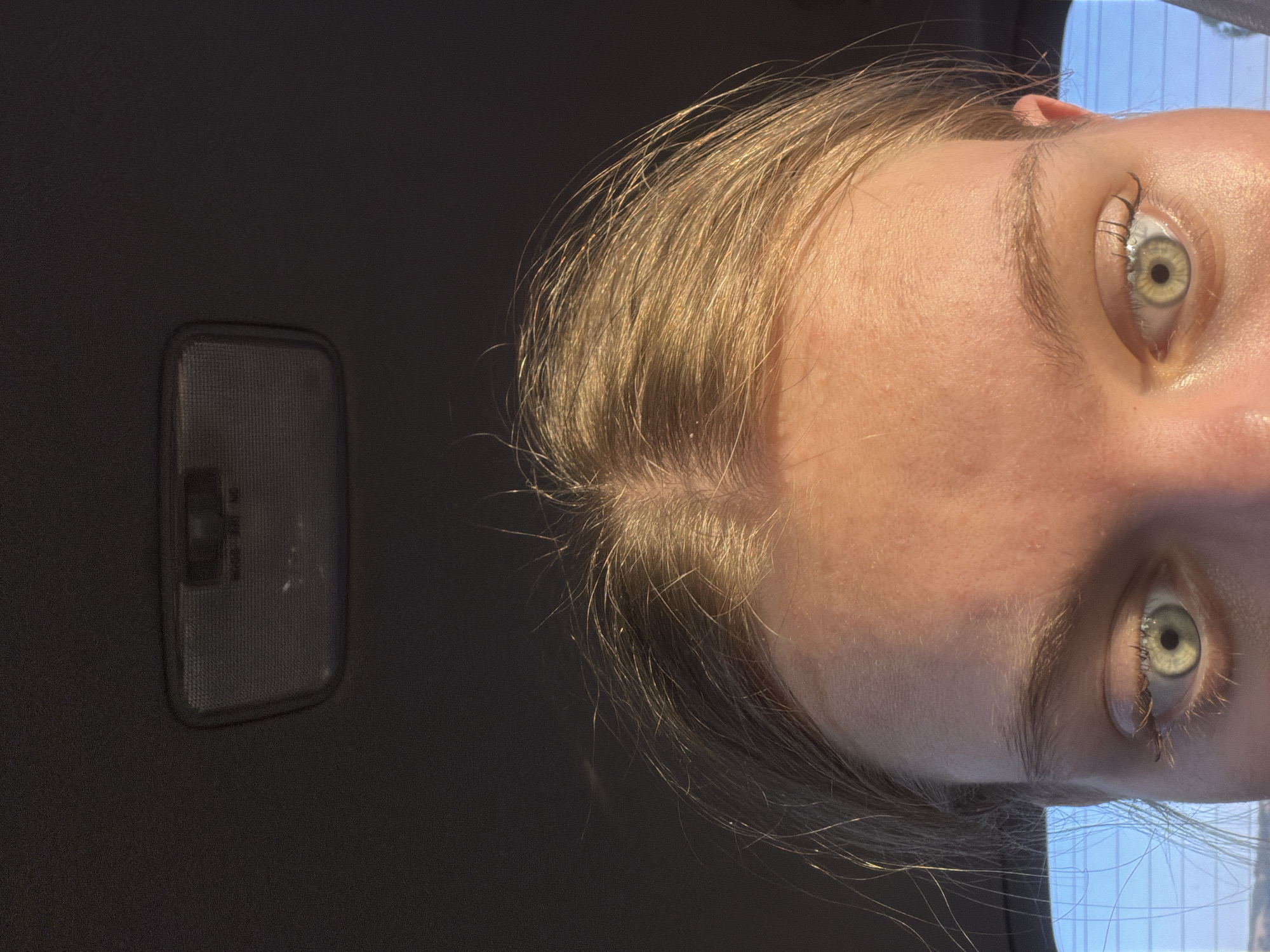 The image shows a close-up, head-on view of a person's face from the eyebrows down. The subject has blonde hair with some strands illuminated by sunlight, indicating a likely outdoor setting or exposure to bright light. Their eyes are light green, and their skin appears fair with some natural texture visible. The upper part of the image shows a dark, textured ceiling with a rectangular light fixture. The background is dark, suggesting an indoor environment such as a car interior, with a faint hint of a blue sky and possibly a window frame visible on the left side, implying the subject is likely in a vehicle. There is no information in the image to place the scene in Belogorki, Russia.