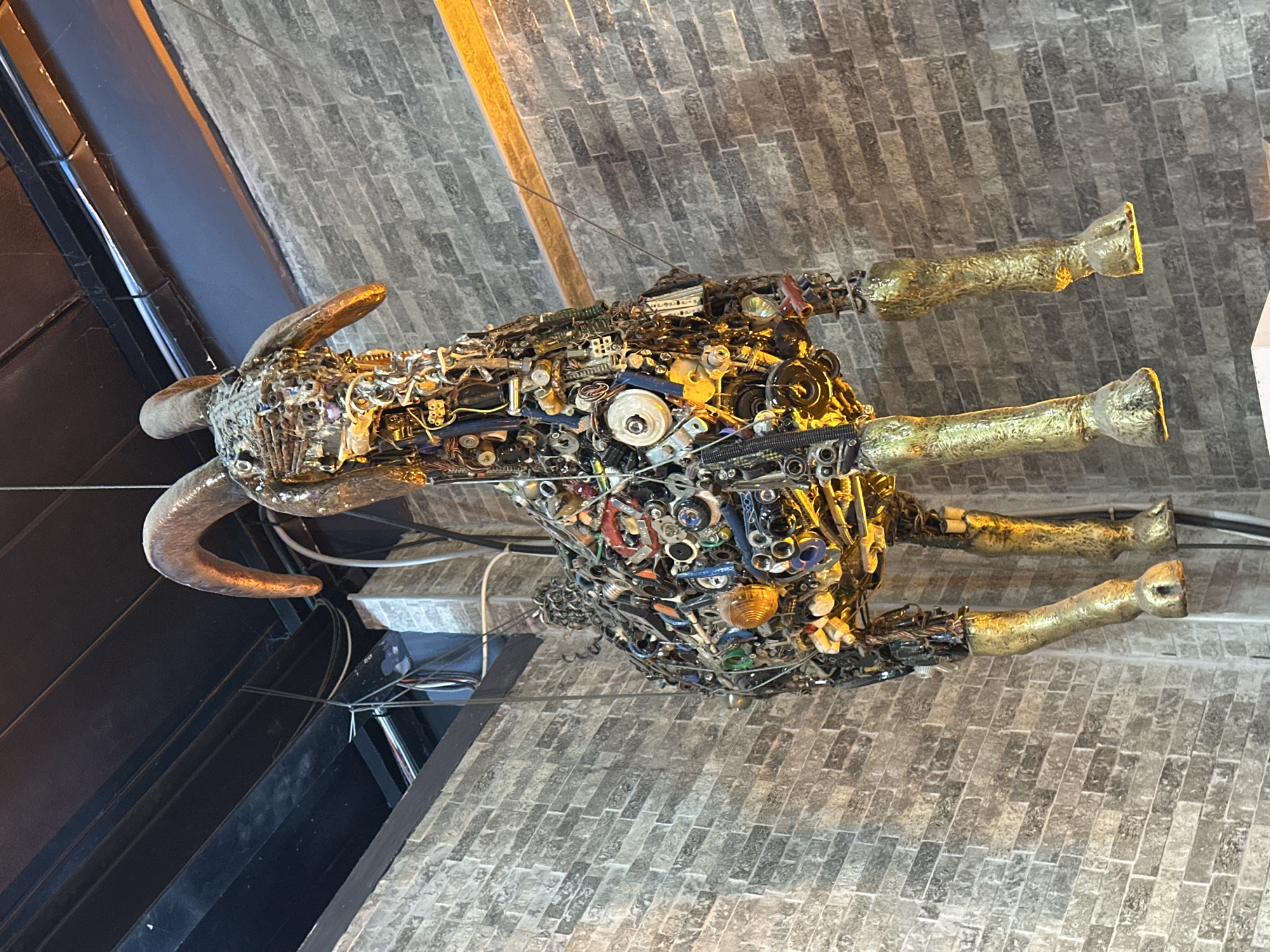 This image shows a sculpture of a goat hanging from the ceiling. The goat is made of various recycled metal parts, including gears, wires, and fasteners. Its horns and legs are a metallic gold color. The sculpture is suspended in front of a brick wall, and overhead, metal beams and electrical wiring are visible. The overall impression is one of an artistic and industrial aesthetic, potentially a decorative piece in a commercial or public space. The location is identified as Altındağ, Turkey.