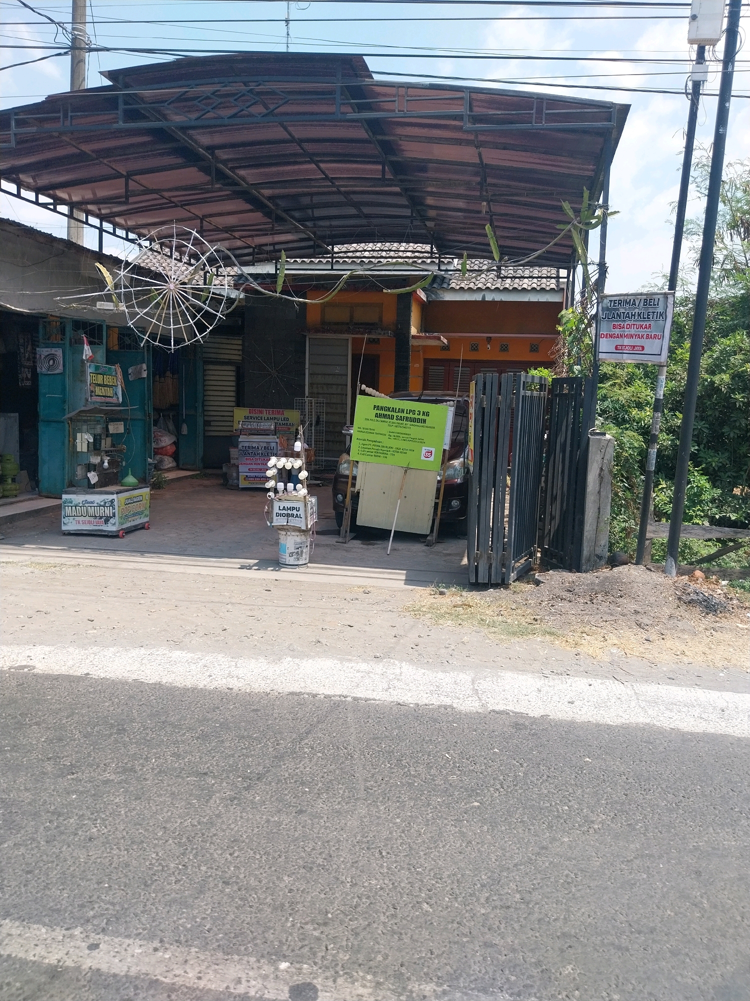 The image captures a street-level view of a small roadside shop in Campur, Indonesia. The shop appears to be an open-air structure with a corrugated metal roof supported by metal framing. Underneath the roof, there is a display of various items, including what seem to be gas cylinders, a small refrigerator or freezer unit labeled "MADU MURNI", and a stand with multiple light bulbs. A sign with Indonesian text, possibly advertising services or products, is visible near the entrance. In the foreground, there's a paved road with a white line marking. The background shows lush green foliage and more of the shop's structure. The overall impression is that of a typical small business operating along a road in a semi-rural or suburban area.