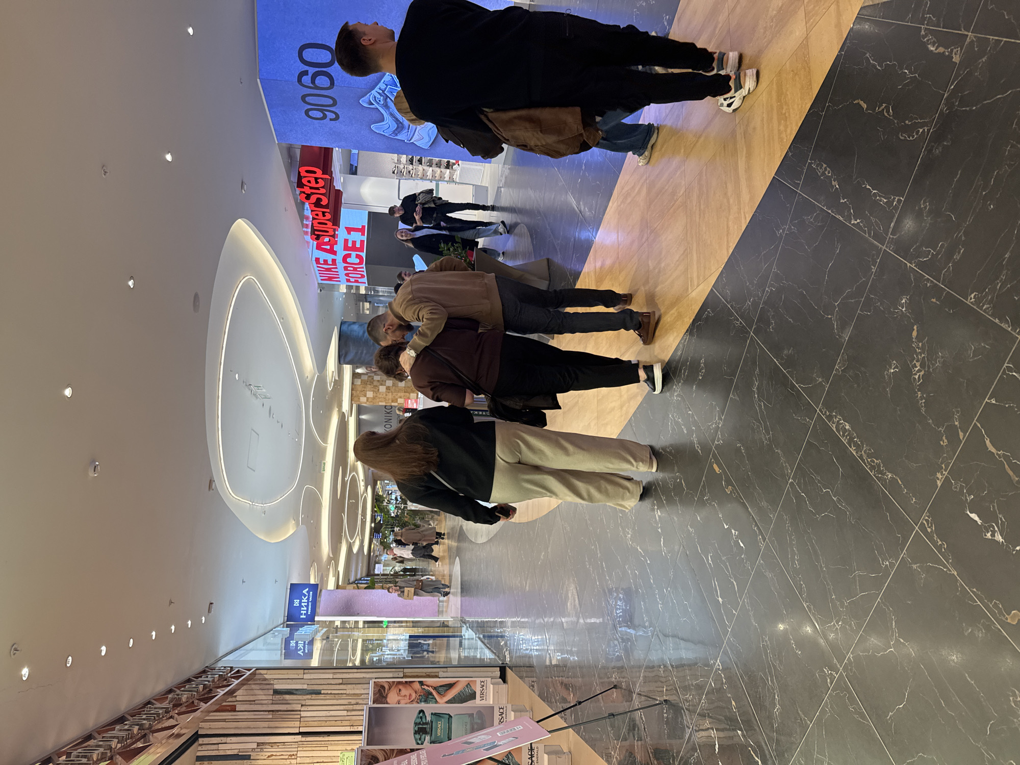 A busy shopping mall in Olginskaya, Russia, features a black and gold tiled floor, a bright white ceiling with recessed lighting, and various retail displays. In the foreground, a display case with cosmetic products is visible. Several people are walking or standing in the mall, some in groups, including a hugging couple. Various advertisements and signs are present.