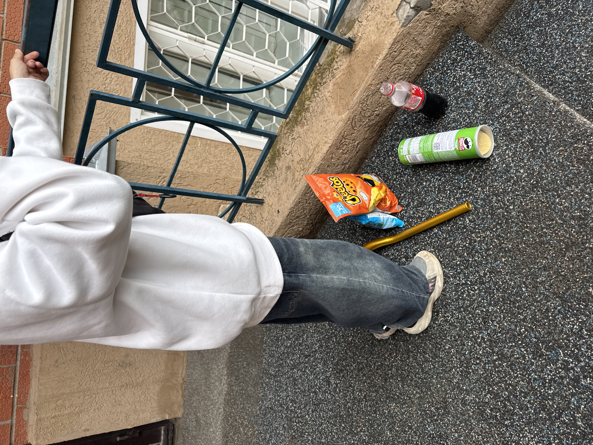 In the city of Chaikovsky, Russia, an outdoor scene captures a person standing on a speckled dark grey aggregate surface. The individual, seen from the waist down and partially from the back, is wearing an oversized white hooded sweatshirt and loose-fitting, faded dark blue denim jeans. Light-colored sneakers with a reflective strip on the heel are visible on their feet. Their right hand rests on a dark teal metal railing with geometric patterns.
On the ground near the person's feet, several items are present: an orange bag of Cheetos snacks, a green open Pringles can containing chips, a clear plastic bottle with a red cap partially filled with a dark liquid (likely a soft drink), and a golden-colored cylindrical object.
The railing extends along a low, tan-colored concrete ledge, behind which a building facade with a light brown stucco texture and a window with white frames and hexagonal-patterned grilles is visible. A red brick wall is partially discernible in the upper background. The scene suggests a momentary pause or a casual outdoor setting on a balcony or walkway.