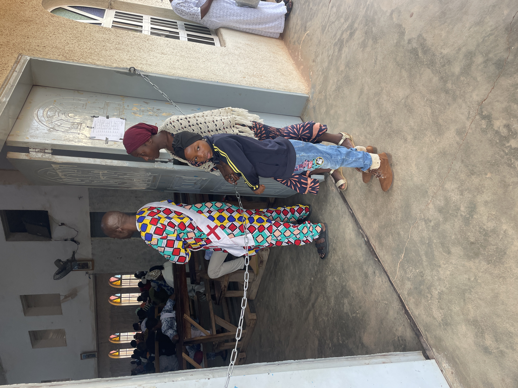 Outside of a building with a gray door secured by a chain, three people stand. Two women, one wearing a red headscarf and the other a black head covering, flank a child wearing jeans, a black jacket, and brown boots. A person in a colorful geometric patterned garment with a red cross stands inside the building, looking outwards. Many people are seated inside the building on wooden benches. The location is possibly Jos, Nigeria.