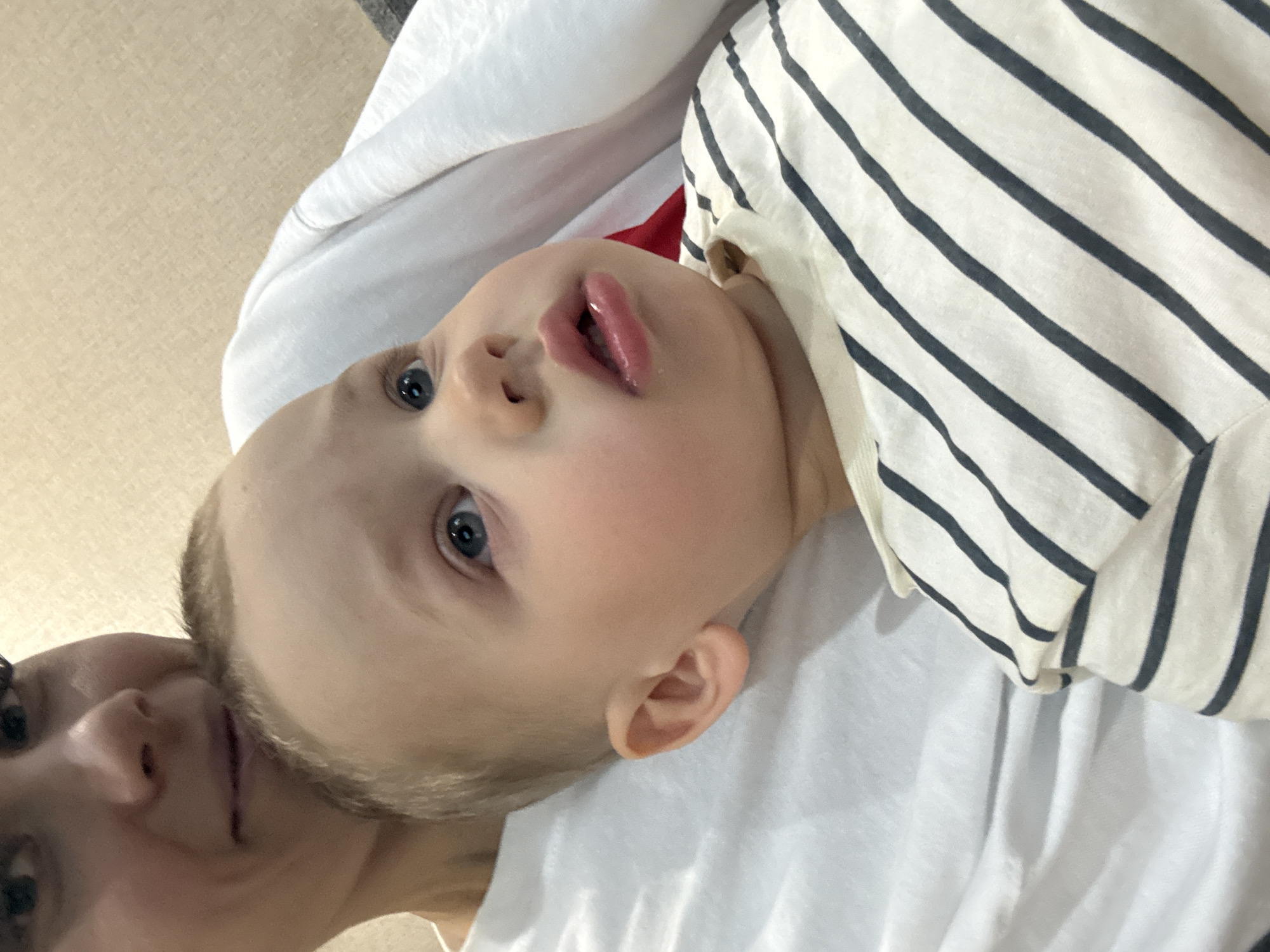 A close-up shot features a light-skinned baby with blue eyes, lying on a white surface. The baby is wearing a white shirt with thin black stripes. A woman's face is partially visible in the bottom left corner of the image. The environment appears to be indoors, possibly a home. There is no indication of location in Rusanovo, Russia, or any specific city.