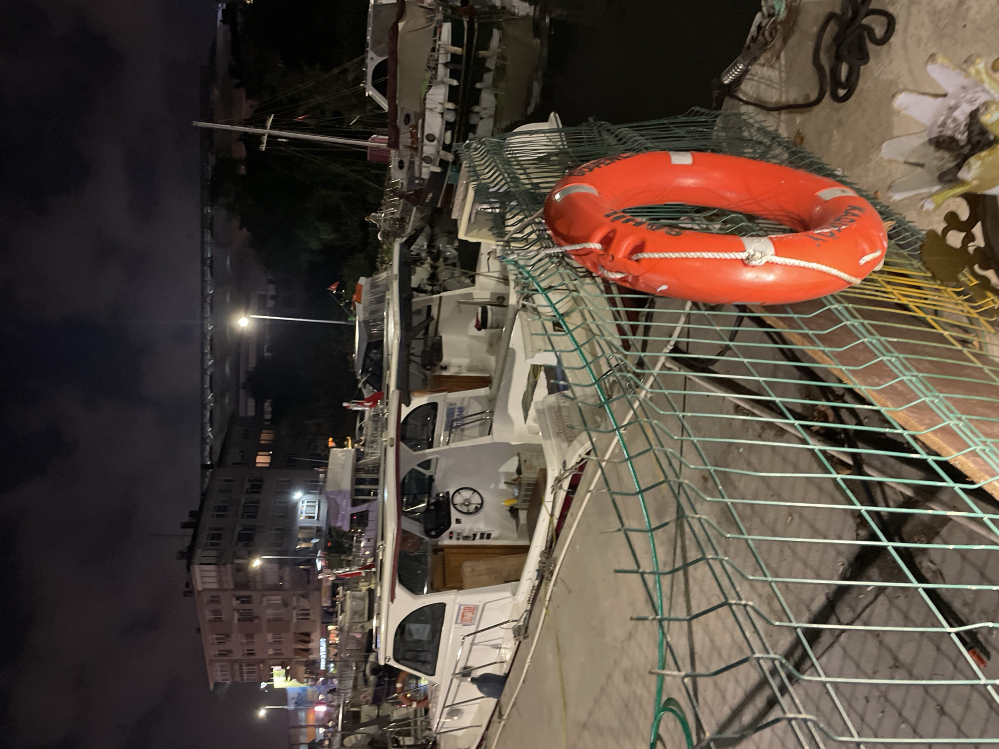 At night, a bright orange life preserver hangs on a green metal fence in the foreground of the image. Behind the fence, several boats are docked in dark water. The closest boat is white with a red stripe and a covered cabin. Further back, other boats of various sizes and designs are visible, some with sails. Across the water, buildings and streetlights suggest an urban environment, consistent with the port area of Kadıköy, Turkey. In the background, a long, elevated structure, likely a bridge or viaduct, spans across the scene. The sky is dark with some clouds, and the overall lighting is dim, illuminated by artificial lights from the city and the boats.