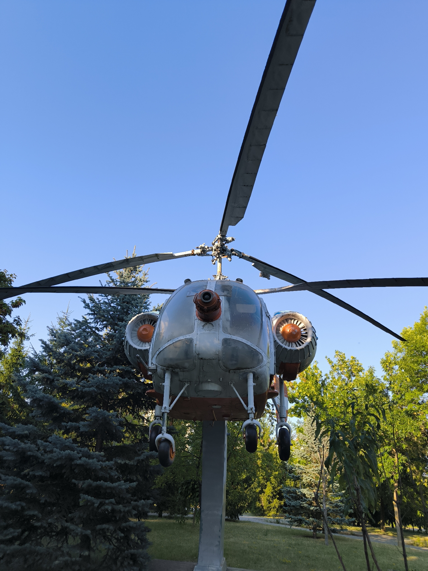 A large, silver helicopter is mounted on a pole in a park with green trees and a clear blue sky. The helicopter is viewed from the front, with its rotor blades extending upwards. Two orange turbine engines are visible on either side of the cockpit. The helicopter's landing gear is down, with two wheels visible. The park appears to be well-maintained, with a grassy lawn and a paved pathway. The scene suggests a sunny day with good visibility. This helicopter is likely an exhibit in the Museum of Military History in Chișinău, Moldova, which often features decommissioned military equipment as outdoor displays.