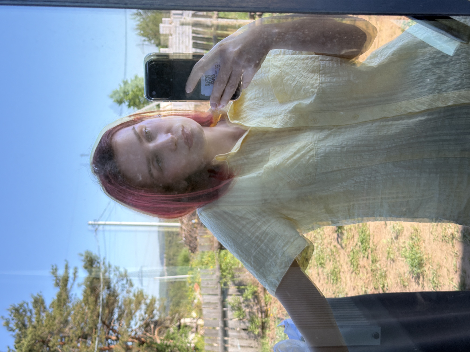 A reddish-haired person in a pale yellow short-sleeved shirt takes a selfie in a mirror or window. The phone is a dark color with a clear case. The background appears to be outdoors, with trees, a blue sky, and what looks like a garden or field visible through the window or in the reflection. There are also power lines and some sort of fencing or constructed object visible. It is not possible to determine if this image was taken in Chaikovsky, Russia without any further data.