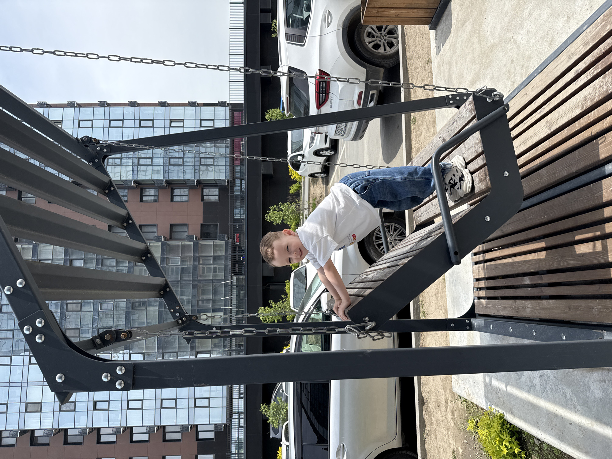 A young boy in a white shirt and blue jeans is climbing on a gray metal and wood bench structure in what appears to be a playground or outdoor recreational area. The boy is facing the camera with a smile. The structure is made of metal with a wooden seating area. Behind the structure are parked white cars, a building with a modern facade featuring multiple windows, and some greenery. The location appears to be urban, possibly a residential area. There is no explicit indication that the image was taken in Vladivostok, Russia.