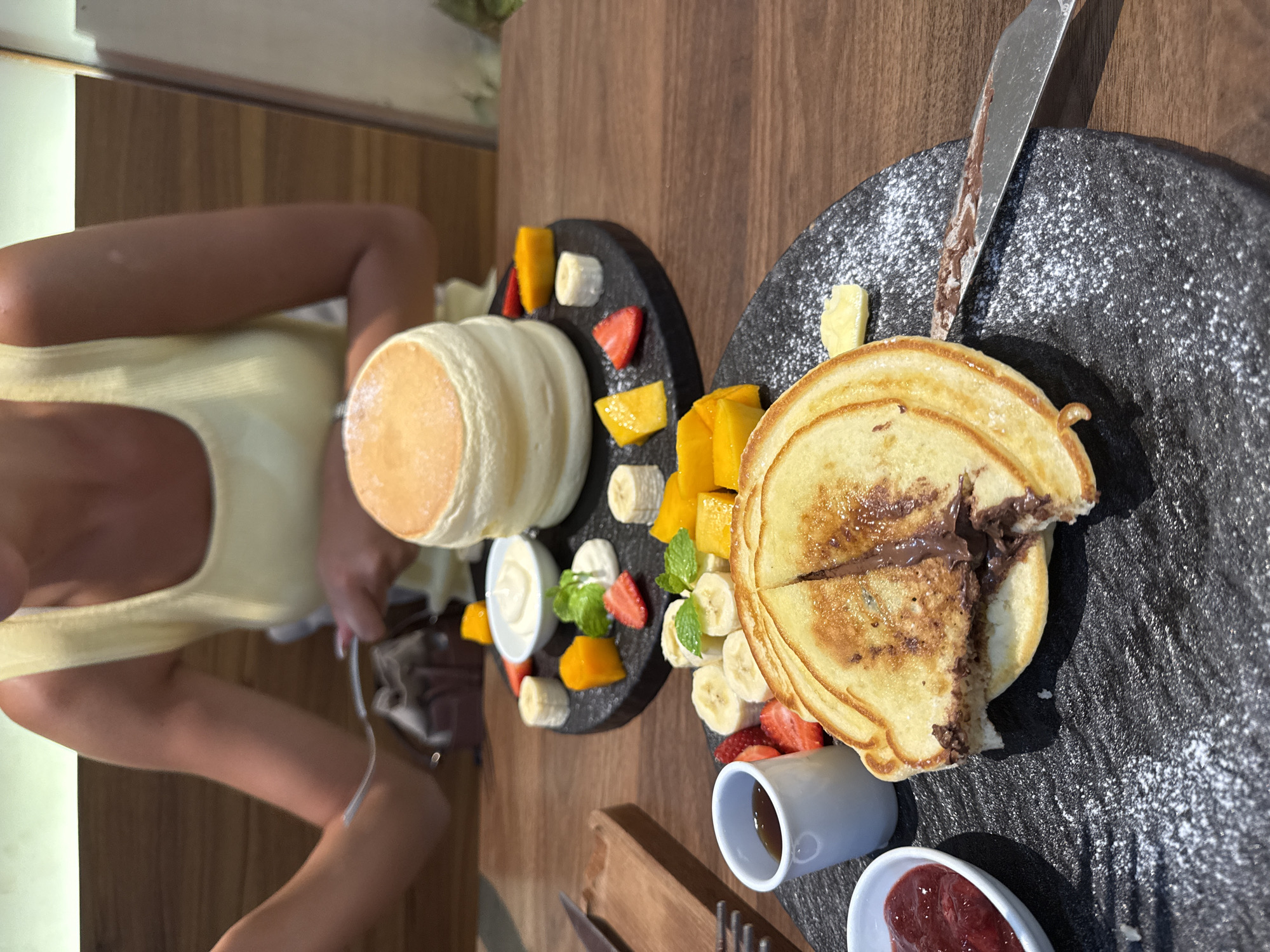 Captured from an elevated perspective, the image showcases a dining experience. Two distinct plates are presented on a wooden table. The left plate features a tall stack of souffle pancakes, accompanied by a white cream, sliced mango, banana, and strawberries. The right plate contains standard pancakes filled with a dark spread, a knife, and a dusting of powdered sugar. Fresh mango, banana slices, and strawberries are also present, along with two small white dishes—one containing a red jam and the other a clear liquid, possibly syrup. The lower portion of a person wearing a light yellow top is visible on the left side of the image, indicating their involvement in the scene. The location is the city of Đà Nẵng, Vietnam.