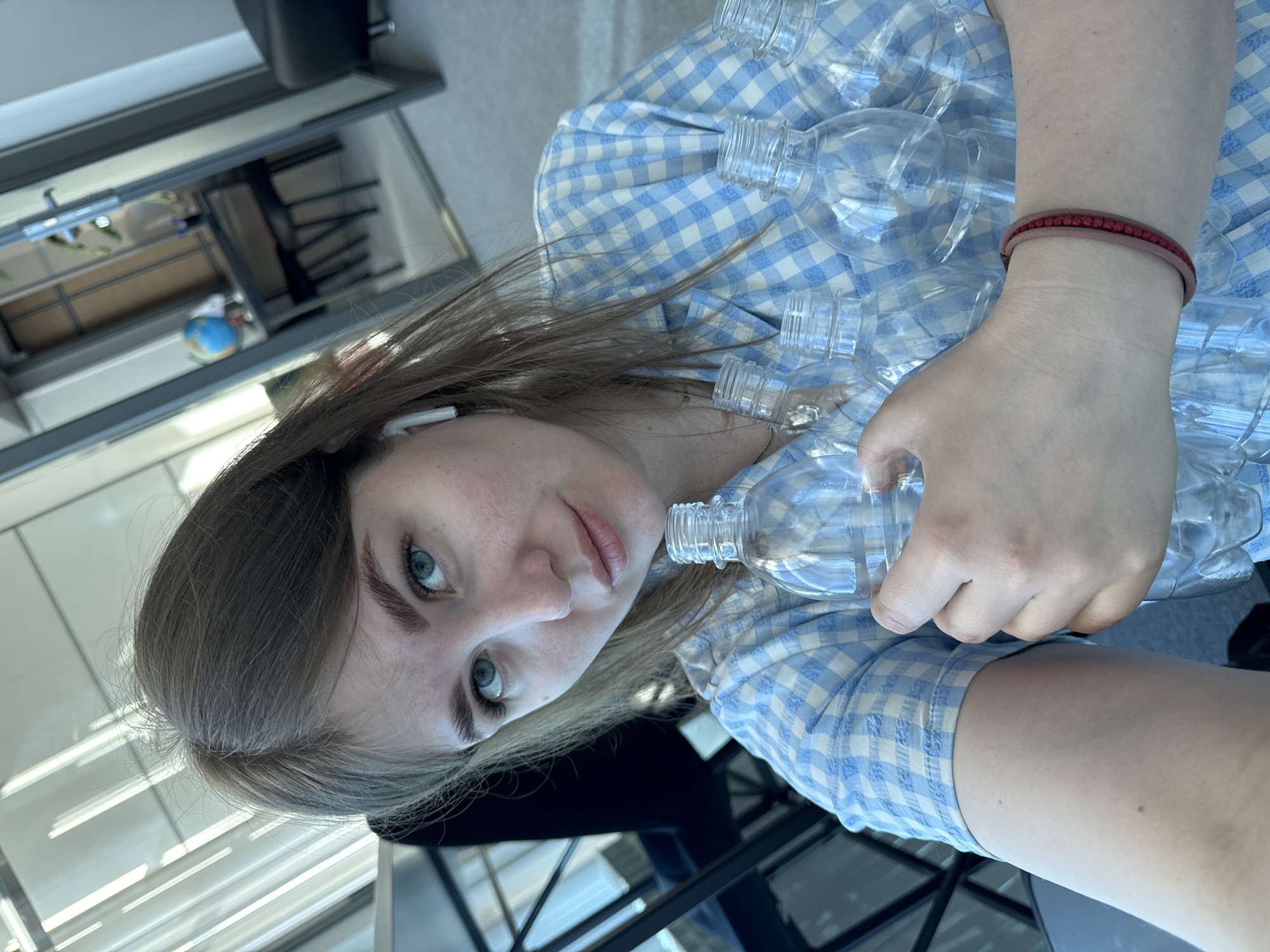 A person, presumably female, is holding multiple empty, clear plastic bottles in their arms. The person has fair skin, brown hair, and blue eyes. An in-ear wireless earbud is visible in their left ear. They are wearing a light blue and white checkered shirt. A red bracelet is visible on their right wrist. In the background, there is what appears to be an interior space, likely an office or waiting area, with chairs and a shelf visible. A miniature globe is on a shelf. The image was captured from a close-up, slightly tilted perspective. The location in the city of Perm, Russia, cannot be verified solely from this image.