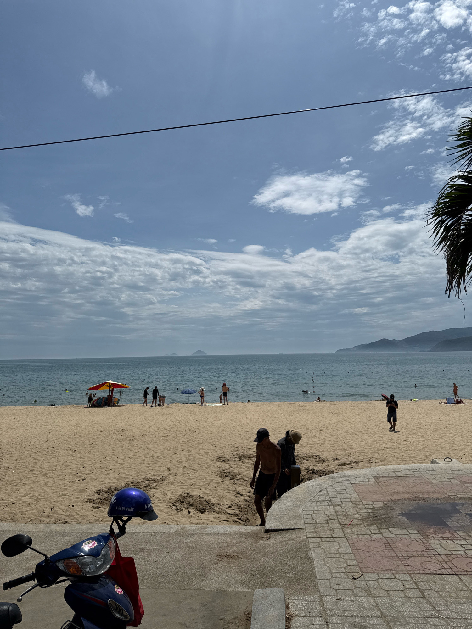 The image depicts a sunny day at Nha Trang beach in Vietnam. In the foreground, a blue and silver scooter is parked, with a blue helmet resting on its handlebars and a red bag hanging from the side. A tiled walkway leads to a sandy beach where several people are visible. Some individuals are walking, while others are lounging under umbrellas or swimming in the ocean. The ocean stretches out to the horizon, meeting a hazy mountainous coastline in the distance. The sky is a mix of blue with scattered white clouds, and a single black wire stretches across the upper portion of the frame. The scene suggests a typical day at a popular tourist destination.