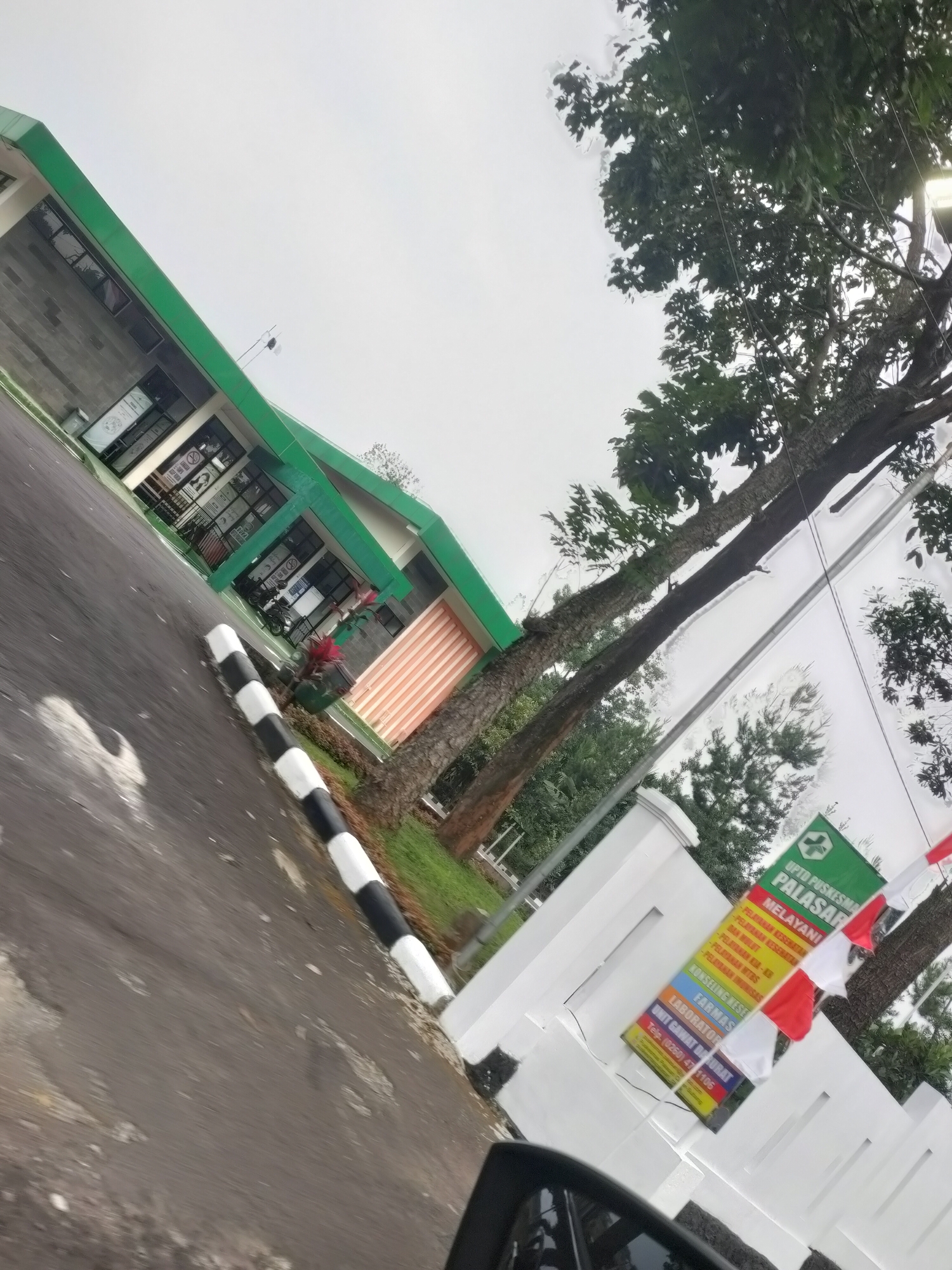 A street-level, slightly tilted outdoor shot shows a modern building with a green roof, likely a public facility, with "UPTD PUSKESMAS PALASARI" text on a green banner, indicating a health center in Palasari, Subang, Indonesia. The banner also lists services such as "PELAYANAN KESEHATAN" (Health Services), "KB DAN MINUM" (Family Planning and Drink), "PELAYANAN KIA-AIR" (Maternal and Child Health - Water), "PELAYANAN IMUNISASI" (Immunization Services), "KONSELING KESEHATAN" (Health Counseling), "FARMASI" (Pharmacy), and "UNIT GAWAT DARURAT" (Emergency Unit), with a contact telephone number. Two Indonesian flags, red and white, are attached to a pole near the banner. A black and white striped bollard is visible on the asphalt road in the foreground, which appears wet and shows signs of wear and tear. Trees with lush green foliage are present in the background, and a portion of a vehicle's side mirror is captured in the lower right corner. The sky is overcast.