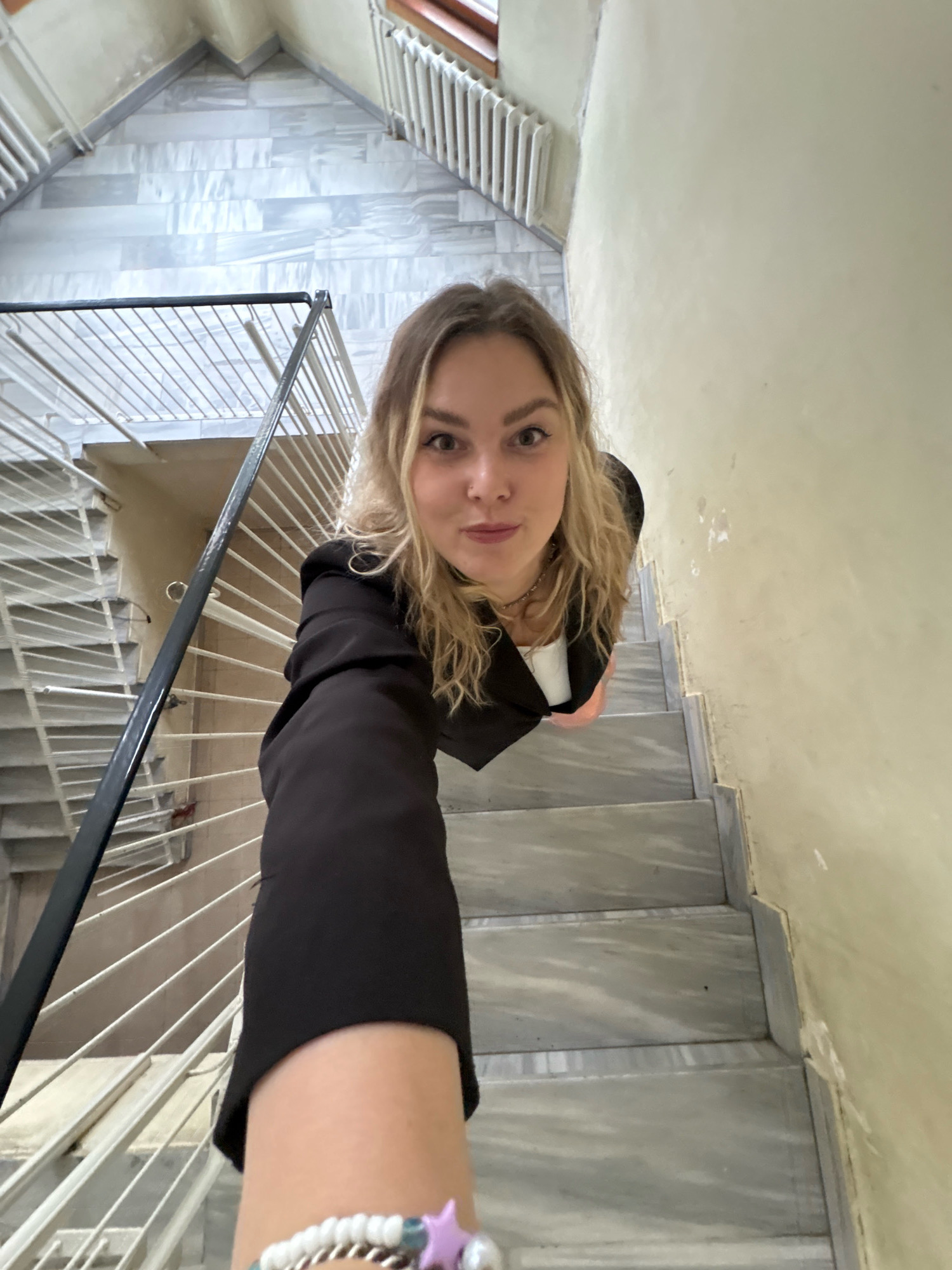 A young Caucasian woman with blonde, wavy hair is taking a selfie from a high angle on a staircase. She is wearing a dark blazer over a white top and a silver necklace. The staircase has marble steps and a light-colored wall with some peeling paint. There is a metal railing to the left of the woman. The background shows more of the staircase, a radiator, and the ceiling. The specific location of Марковский, Russia, is not visible in the image.