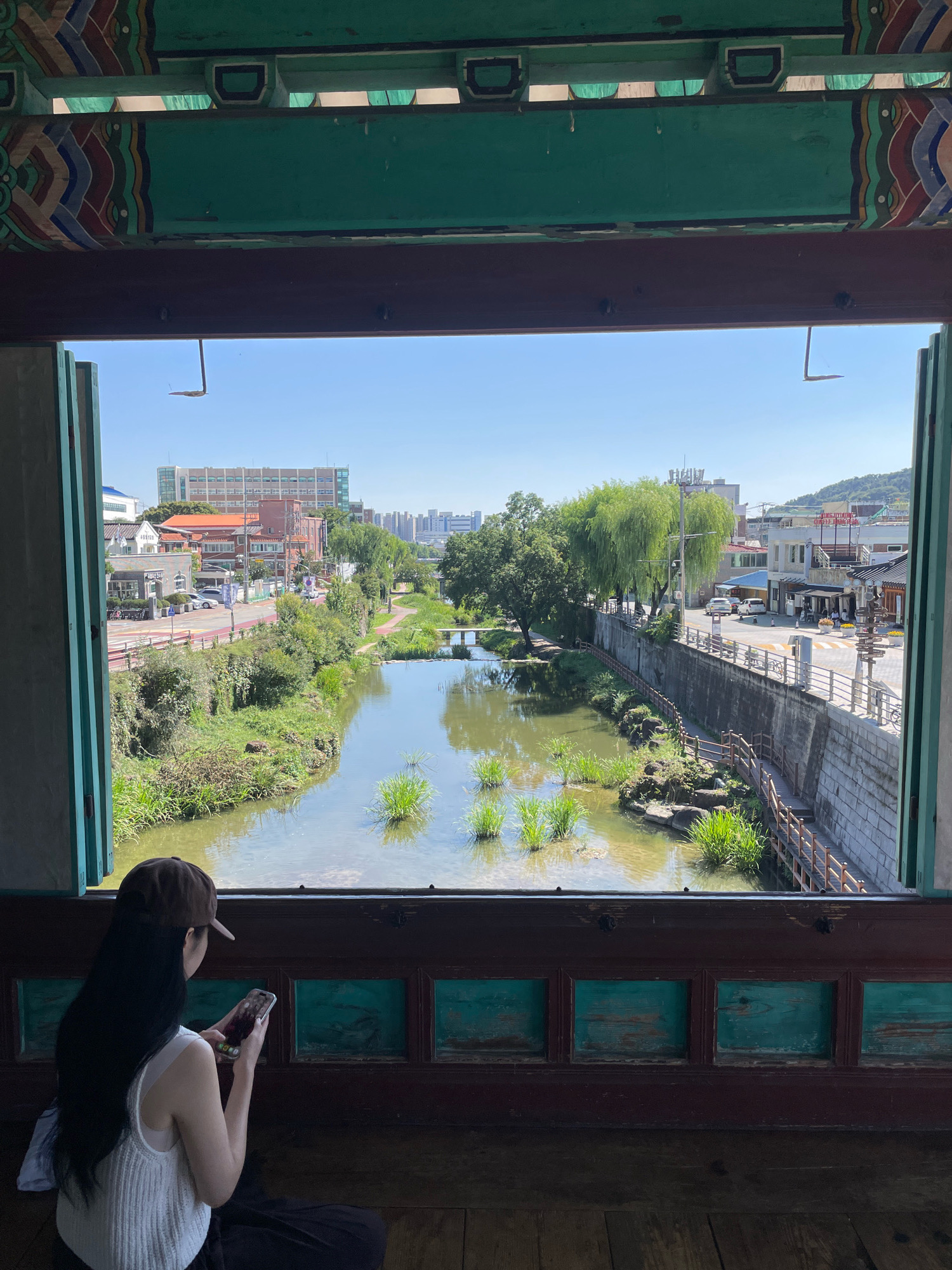 This is a photograph taken from within a traditional Korean structure, looking out a window at a canal in Suwon-si, South Korea. The structure's interior features a dark wooden frame with decorative green panels and intricately painted beams. A person, a woman wearing a white tank top and a brown baseball cap, is seated on the floor in the foreground, facing away from the camera. She is holding a smartphone in her hands, with her long dark hair falling over her shoulders.
The view outside the window shows a canal with clear water and green aquatic plants. The canal banks are lined with lush vegetation, and a wooden walkway with railings follows the contour of the canal on the right side. Beyond the canal, a street is visible with cars, buildings, and pedestrians. The architecture of the buildings suggests a mixed urban and traditional setting. In the background, there are taller modern buildings and hills. The sky is clear and blue, indicating a sunny day. The composition frames the external scene within the ornate interior of the structure, creating a contrast between the man-made elements and the natural environment.
