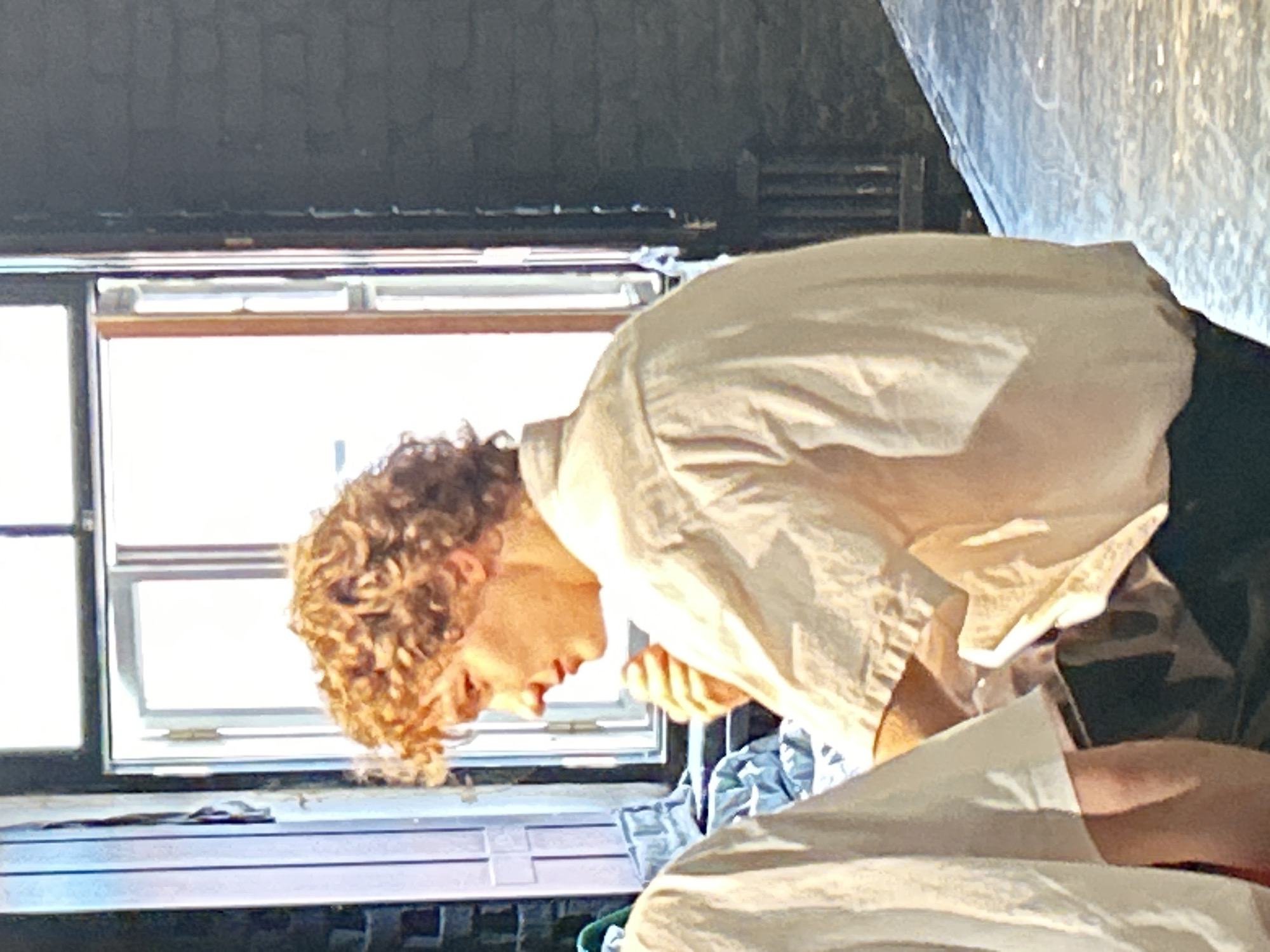A person with curly blonde hair is bent over in front of a window. They are wearing a light-colored, possibly white, collared shirt. The window has thin wooden frames and appears to be divided into multiple panes. Outside the window, the scene is very bright, suggesting daylight. The interior surroundings are dimly lit, with dark walls and what looks like a cluttered surface below the window. The person's face is in profile, and their expression is difficult to discern due to the angle and lighting, but they seem focused on something below. There is a hint of fabric or clothing in the foreground, in a light color similar to the shirt. The overall impression is of a candid moment captured indoors, possibly in a rustic or unfinished setting.