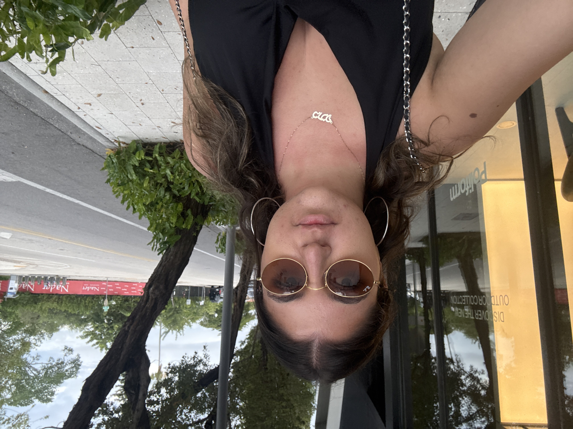 The image is an upside-down selfie of a woman wearing sunglasses, hoop earrings, and a necklace with the word "Chloe" on it. She is wearing a black V-neck top. The background shows a street scene with trees, a sidewalk, and a building with a sign that reads "DISCOVER THE NEW OUTDOOR COLLECTION." A banner reading "Nader" can be seen on the other side of the street. The location is likely Miami, United States of America.
