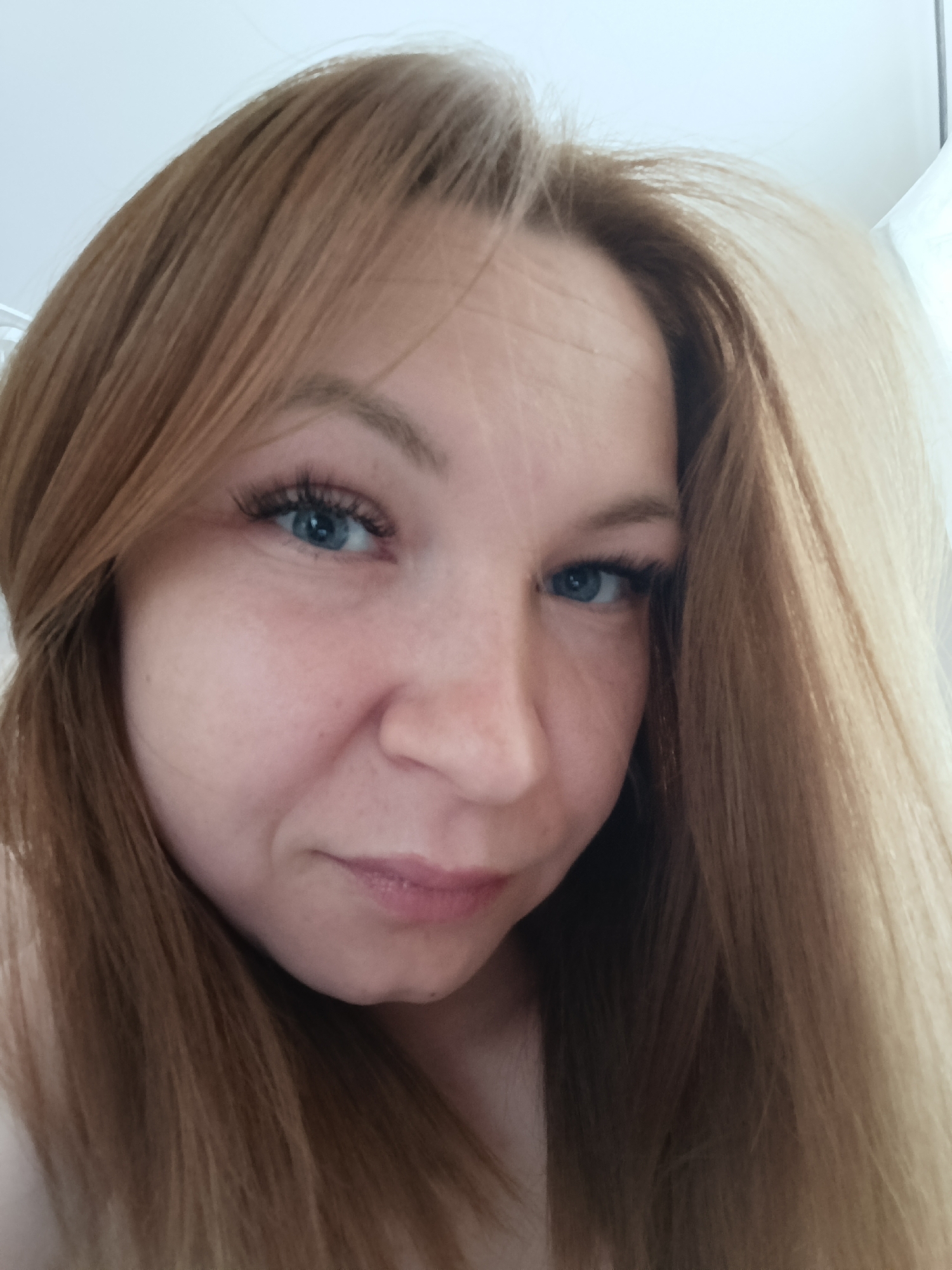 A close-up selfie of a woman with fair skin, blue eyes, and light brown to blonde hair. She is looking at the camera. Her hair is straight and long, partially covering her shoulder. False eyelashes are visible. The background appears to be a bright, neutral color, possibly a wall. No other objects or subjects are visible. The image provides insufficient details to determine a specific location in Chaikovsky, Russia.