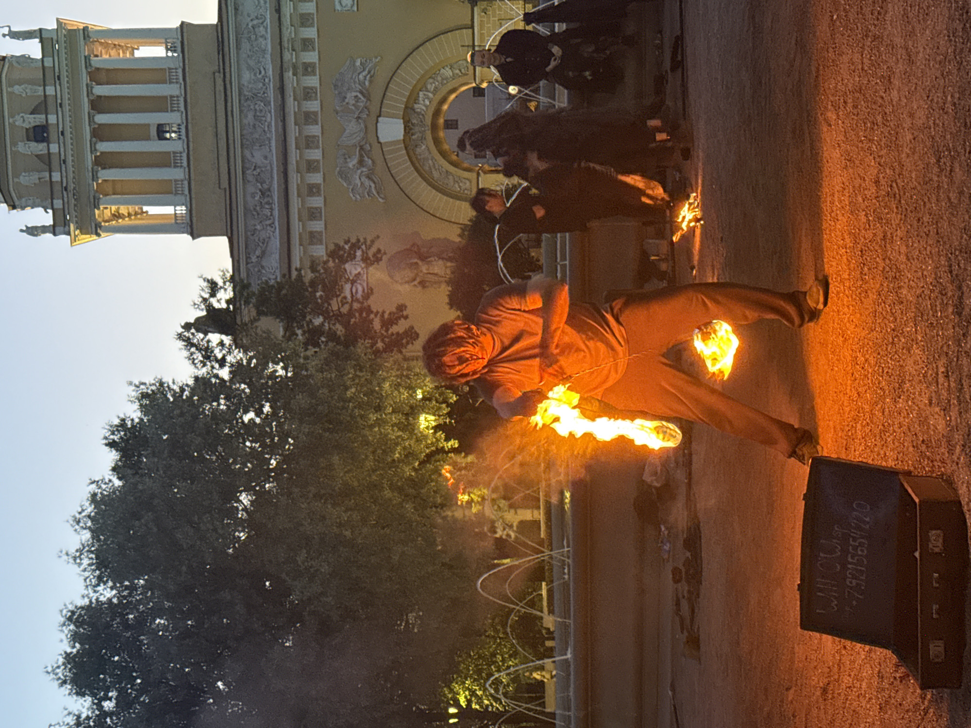 In Saint Petersburg, Russia, a fire performance is taking place near the Admiralty building. A man with dreadlocks is actively performing with fire, holding a flaming object in his right hand and mid-spin. He is wearing a gray t-shirt and loose-fitting tan pants. In the foreground, an open, dark-colored briefcase displays a handwritten inscription and a phone number. Behind the performer, a fountain with arched water jets is visible, with several individuals dressed in dark clothing standing near it. The background is dominated by the ornate architecture of the Admiralty building, featuring classical columns and sculptures, bathed in the warm glow of streetlights and the setting sun. Lush trees frame the scene, with the sky above showing a clear, pale blue hue. The ground is covered in gravel.