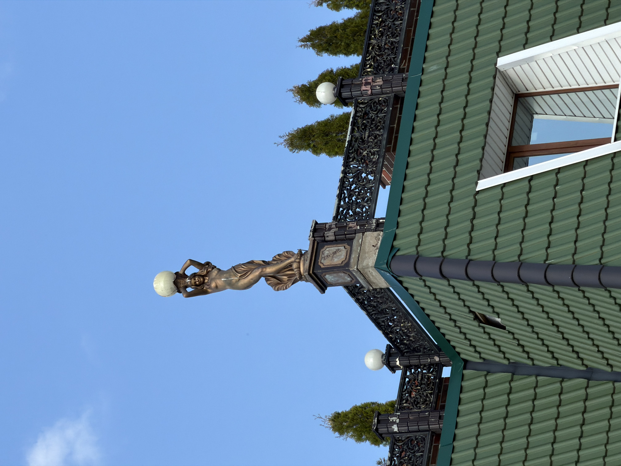 The image shows a building detail in Vsevolozhsk, Russia. A decorative bronze female figure holding a globe is positioned atop a dark wrought-iron railing that runs along the edge of a green tiled roof. There are small evergreen trees planted along the railing. A window with a white frame is visible on the roof. The background is a clear blue sky.