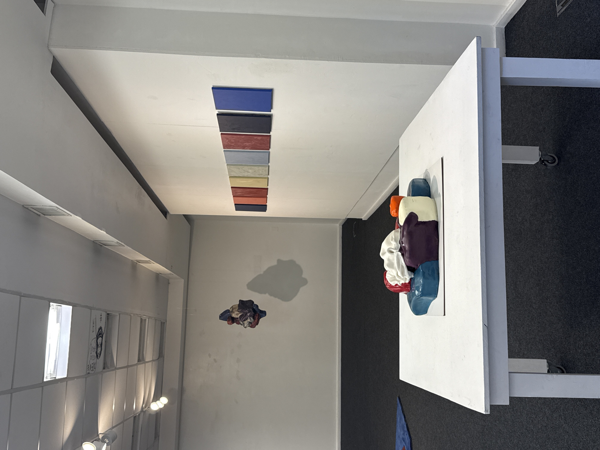 The image captures an interior view of an art gallery or exhibition space in Buenos Aires, Argentina. The room features predominantly white walls and ceiling, contrasted with dark grey carpeting.
In the foreground, a large, white rectangular display table holds a prominent, colorful, abstract sculpture. This artwork is composed of several rounded, organic forms in various hues, including white, purple, blue, red, and orange, possibly suggesting draped fabric or intertwined elements.
On the ceiling, a series of ten rectangular panels are arranged in a horizontal line. These panels display distinct solid colors, ranging from dark blue and black to red-brown, light blue, grey, cream, yellow, and orange-red, creating a vibrant visual strip.
Further into the room, on a white wall to the left, a smaller, similarly abstract and colorful sculpture is mounted, casting a soft shadow. In the background, also on the left, a section of the wall features a grid-like design with embedded lights. On this wall, a piece of paper or poster is visible, displaying an abstract drawing of a face and some partially legible text that appears to include "DEVE" and other symbols.
The scene portrays a well-lit, minimalist environment, characteristic of an art exhibition, with artificial lighting illuminating the artworks. No individuals are visible, allowing the focus to remain on the art pieces and the exhibition layout.