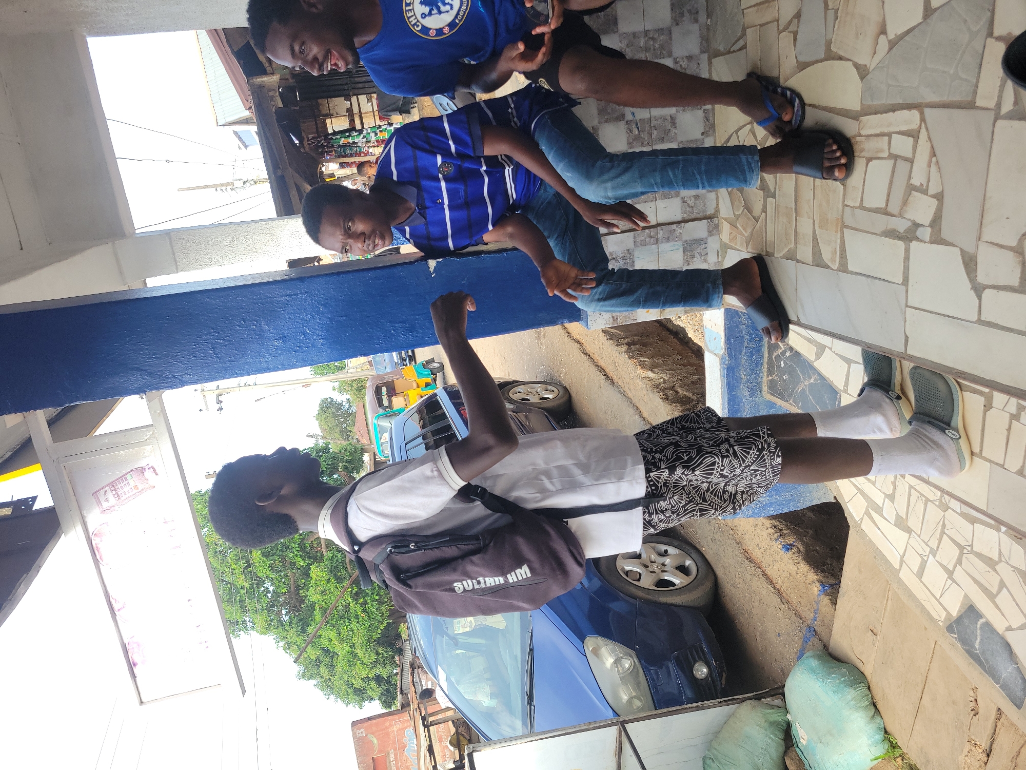 Three people are standing on a tiled sidewalk in the city of Unknown, Unknown. The first person is wearing a white shirt and black and white patterned shorts, white socks, and grey shoes. They are also wearing a backpack. The second person is wearing a blue and white striped shirt and jeans, with sandals. The third person is wearing a blue Chelsea shirt and dark-colored shorts, also with sandals. There are cars parked on the street, and green bags are on the sidewalk. A blue structural beam runs along the top of the frame. A building is visible in the background.