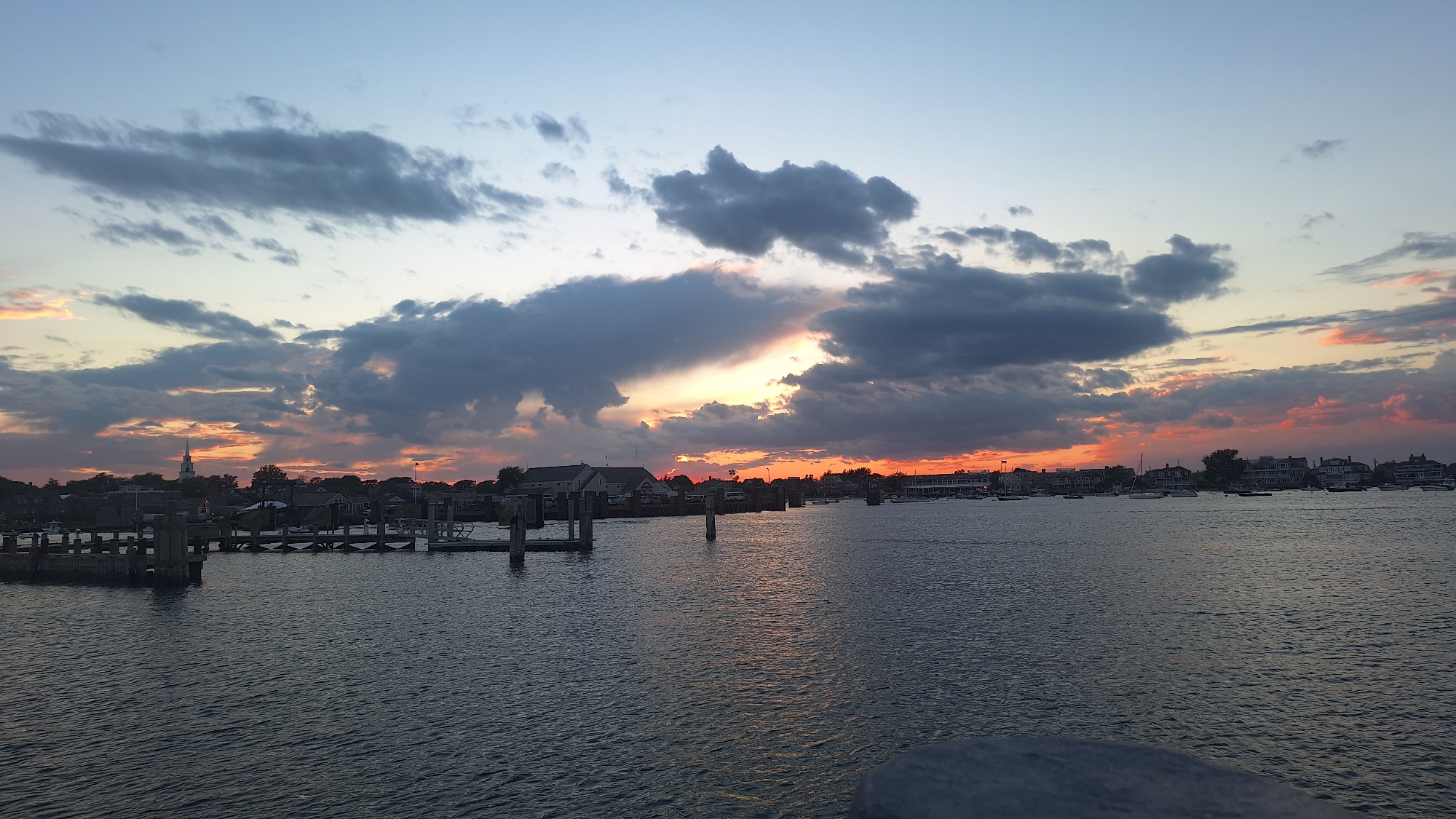 This is a wide, horizontal, eye-level photograph of a harbor at sunset, likely in Nantucket, Massachusetts, given the architecture and waterfront presence. The sky is filled with dramatic clouds, ranging from deep grays to soft pinks and oranges, illuminated by the setting sun. The sun itself is partially obscured by clouds, creating bright streaks of light.
In the foreground, the calm surface of the water reflects the colors of the sky and the silhouettes of boats. A wooden dock with pilings is visible on the left side of the frame, suggesting a harbor or marina. Beyond the water, a line of buildings with a distinctly coastal New England architectural style stretches along the waterfront. A prominent white church steeple is visible among the buildings on the left, further hinting at a New England coastal town. Numerous sailboats and other vessels are anchored in the harbor, adding to the serene atmosphere. The overall impression is one of a peaceful evening in a picturesque harbor.