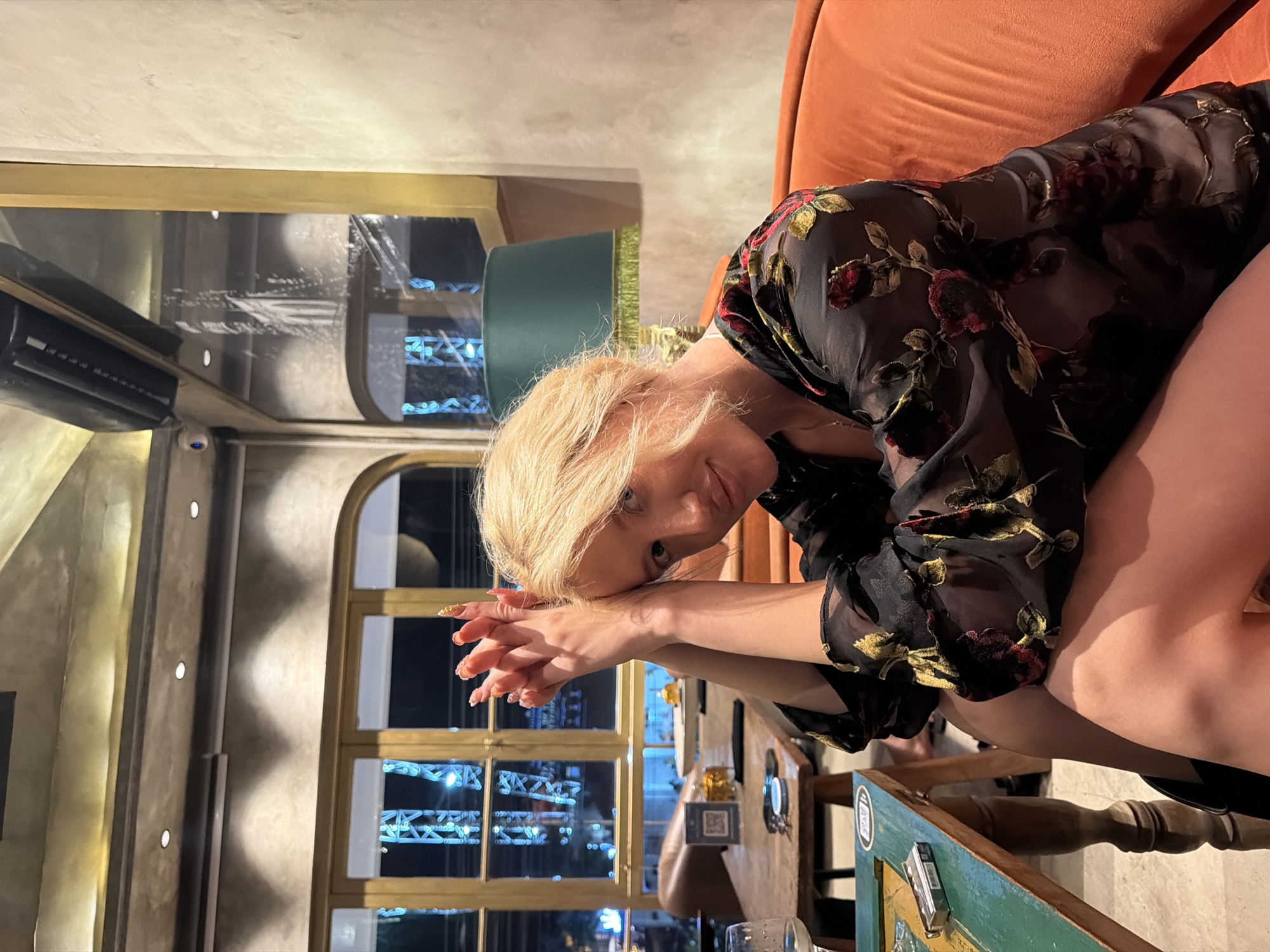 A blonde woman is seated on an orange couch in a dimly lit indoor restaurant. She is leaning forward with her hands clasped together in front of her face. She is wearing a sheer black dress with a floral pattern of red roses and green leaves. The background shows a wooden table with a package of cigarettes and a glass of white wine, and large windows with a view of city lights. The restaurant appears to be located in North Kuta, Indonesia.