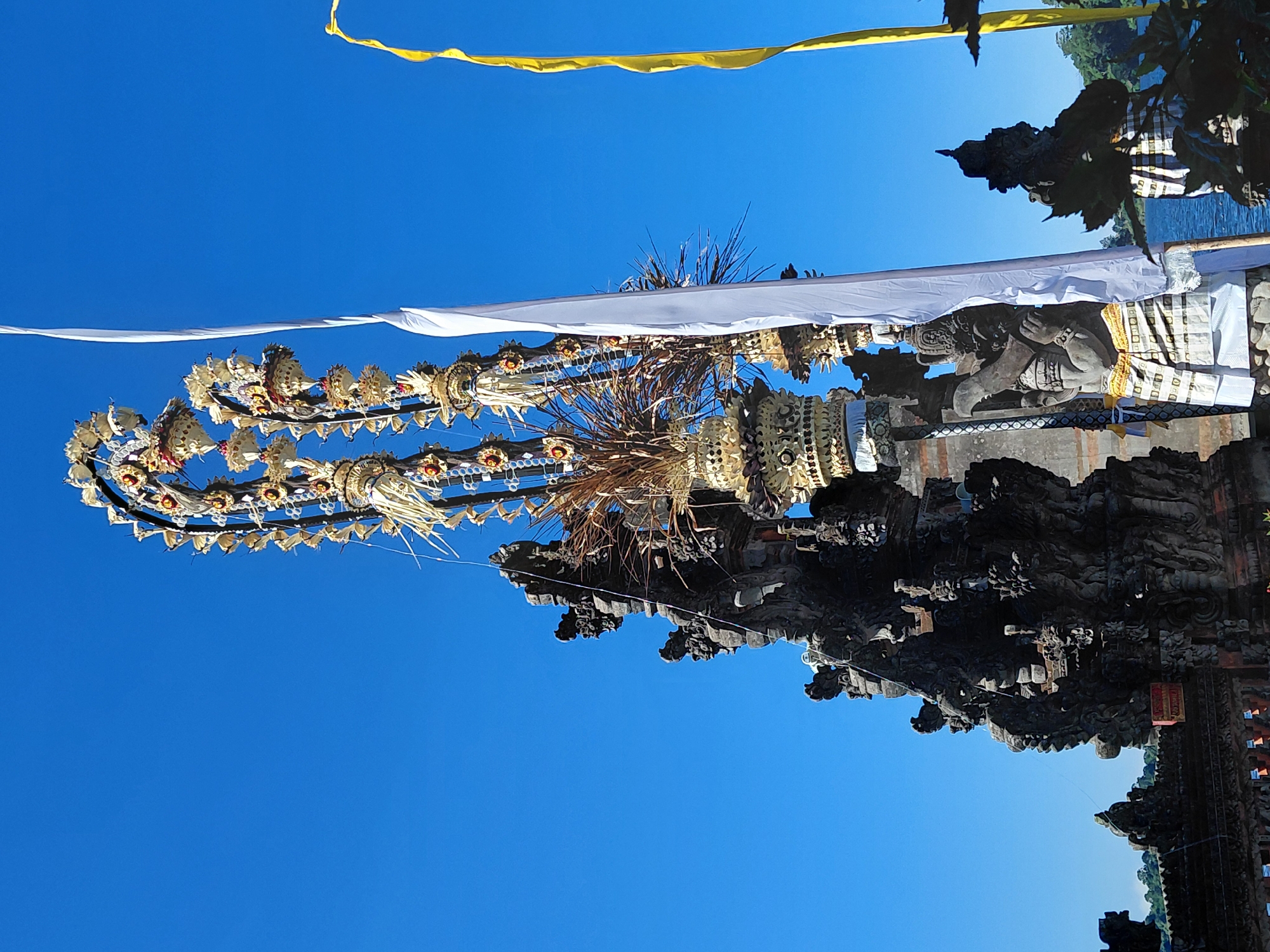 The image depicts an ornate, tall Balinese temple structure with intricate stone carvings and decorative elements, set against a clear blue sky. Dominating the foreground is a tall, multi-tiered structure adorned with golden and white embellishments, resembling offerings or ceremonial decorations. A long white fabric flag hangs vertically on the right side of this structure, and a yellow flag is visible further to the right. The temple itself is constructed from dark stone, with detailed sculptures of mythical figures and architectural patterns. In the background, a body of water, likely a lake, is visible, bordered by lush green trees. The overall scene suggests a religious or cultural site in a tropical environment. This specific temple is likely Pura Ulun Danu Batur, located in the village of Batur, Bangli Regency, near Baturiti, Bali, Indonesia.